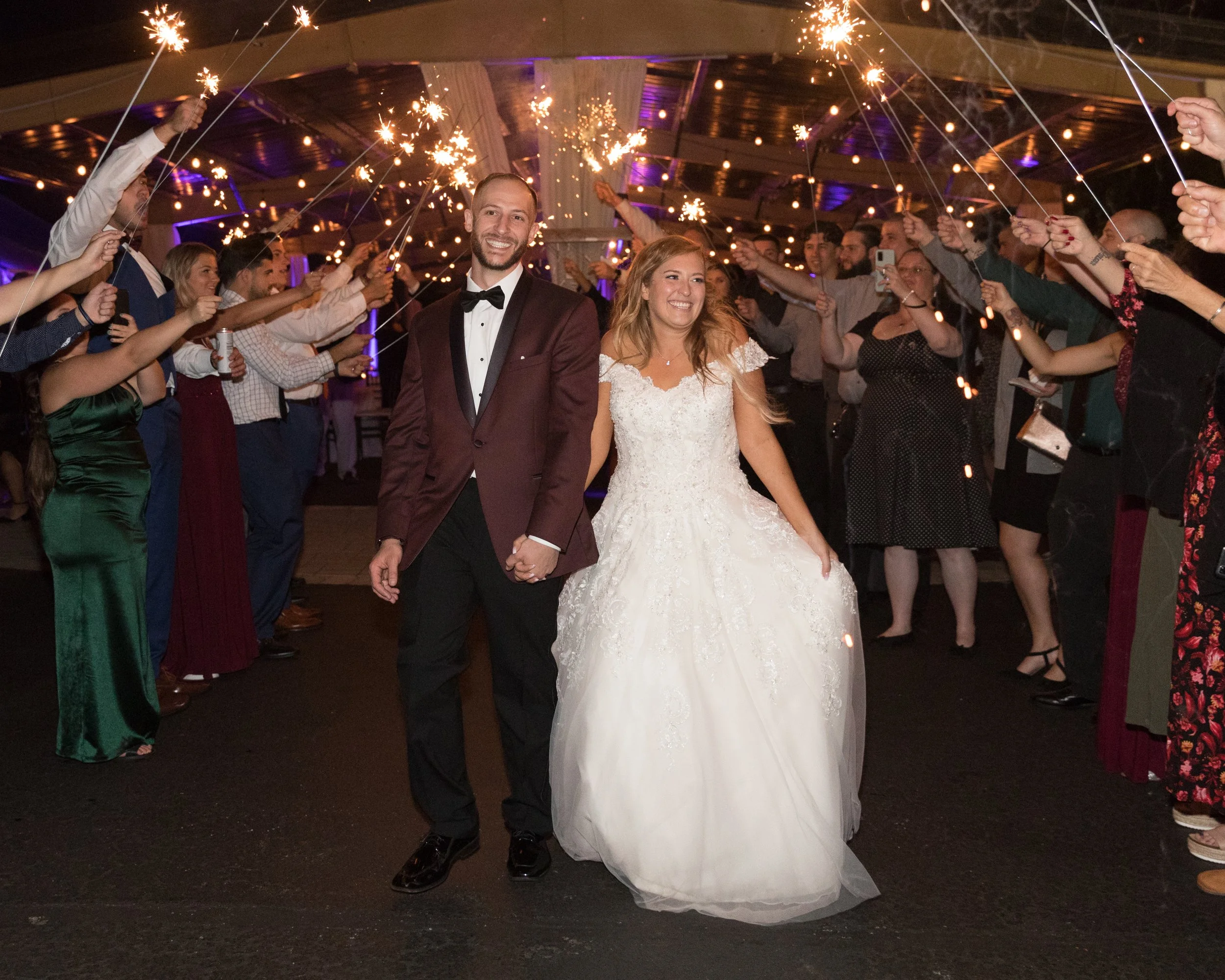 Bride and groom walking under sparklers held by guests in formal attire, nighttime wedding exit.