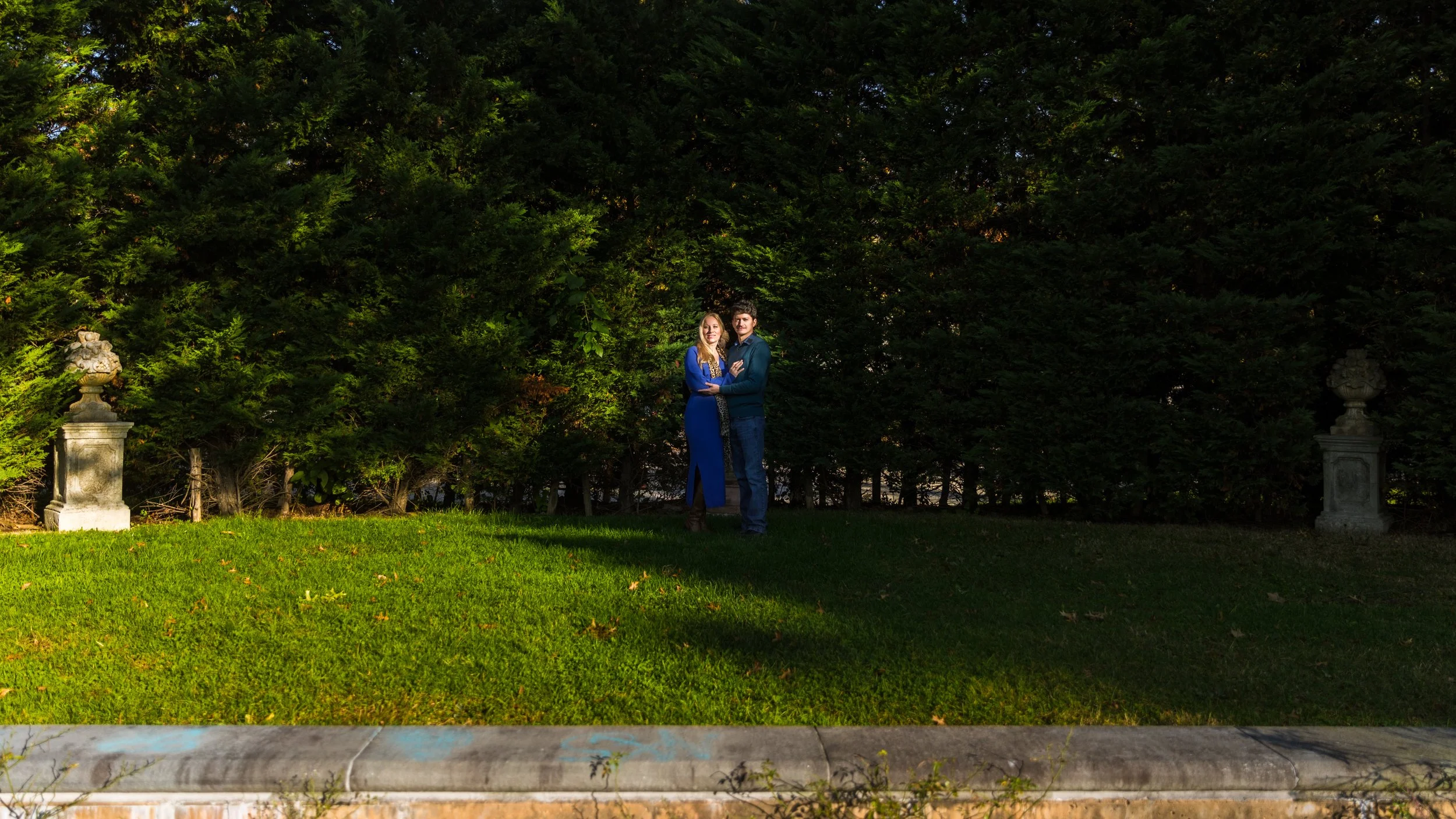 A couple standing on a lawn surrounded by dense green shrubs with decorative stone urns on either side.