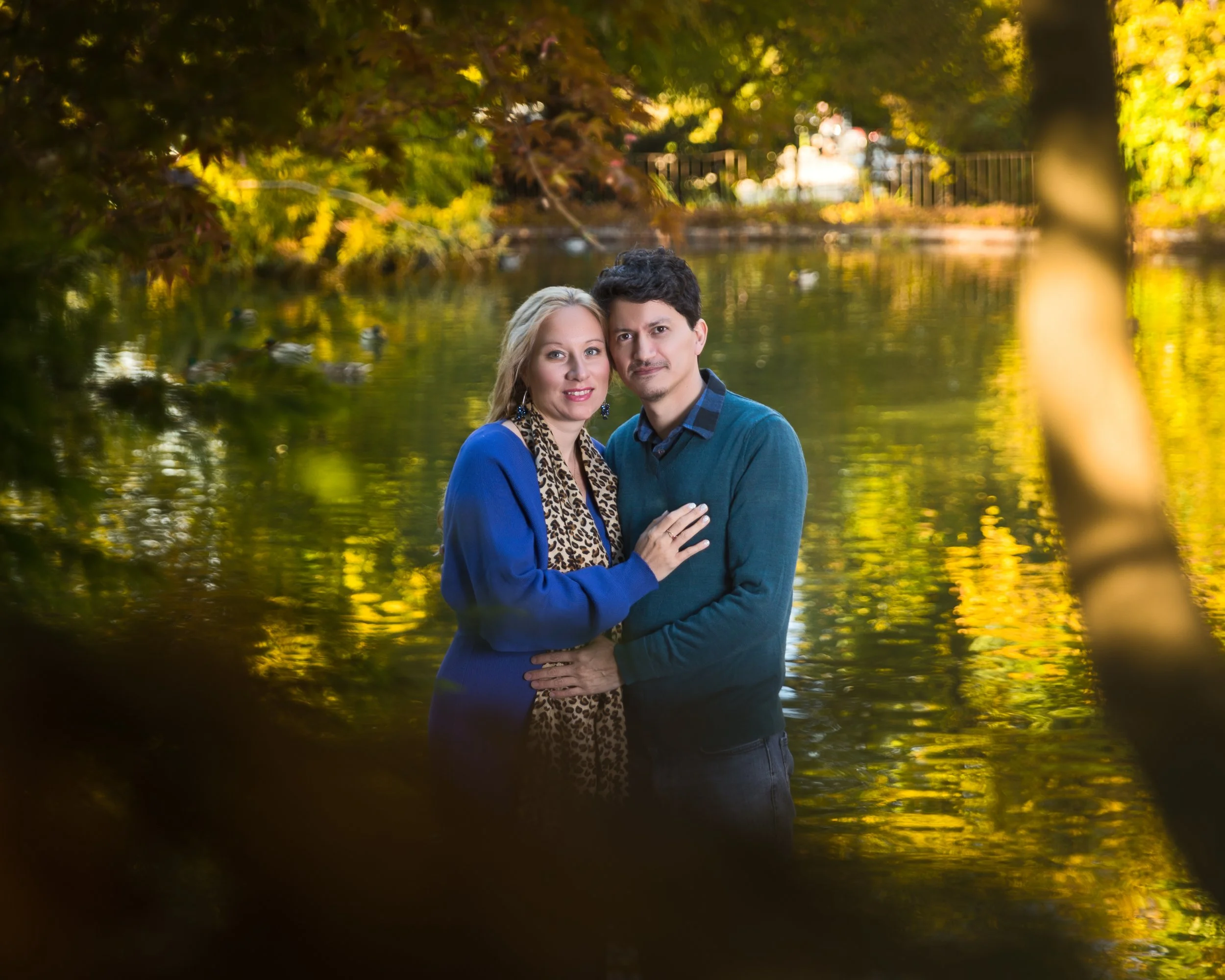 A couple standing close together by a serene lake surrounded by trees with autumn leaves and ducks swimming in the background.