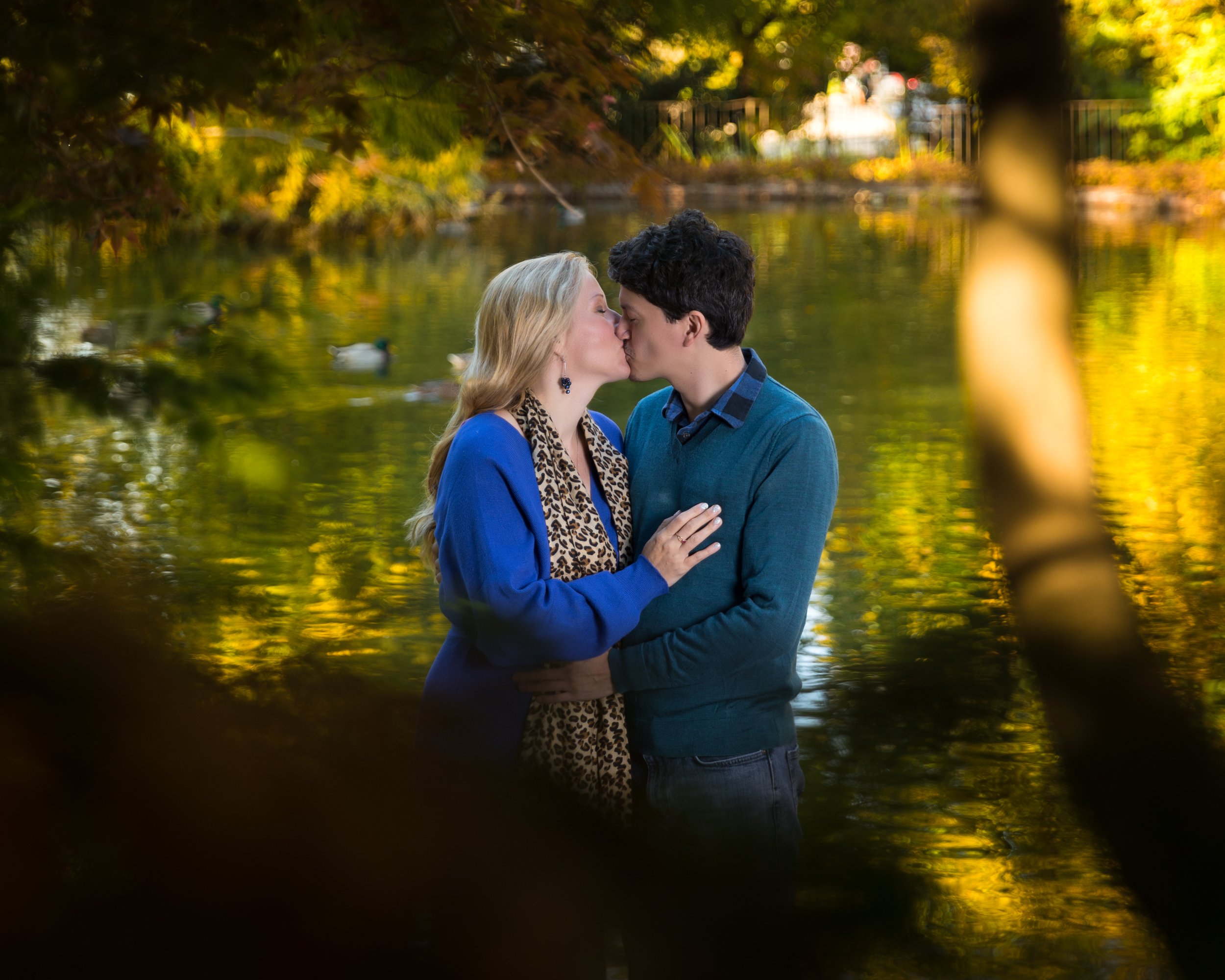 A couple kissing by a serene pond surrounded by autumn trees, with ducks swimming in the background.
