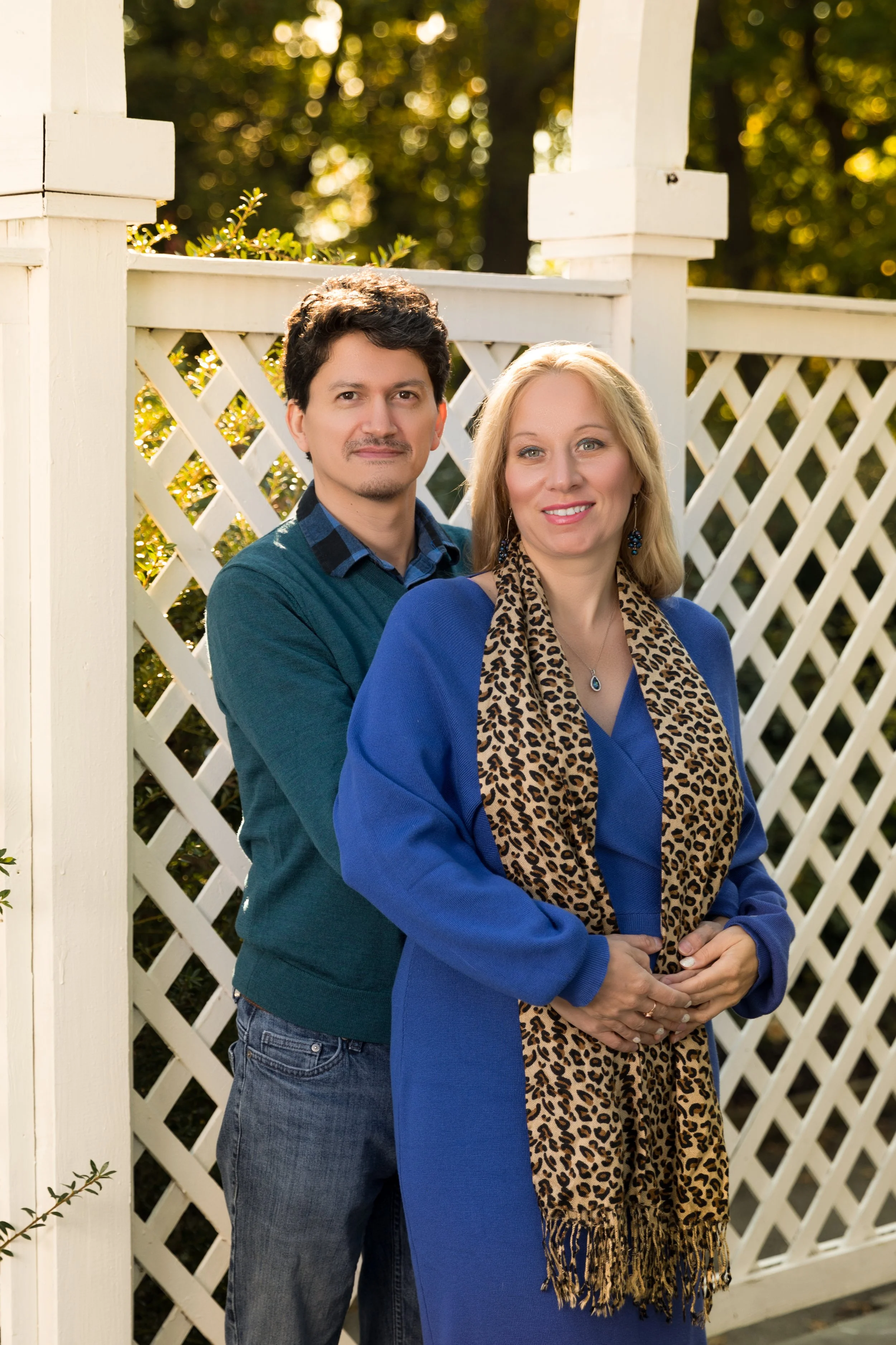 A couple posing for a photo in front of a white lattice garden structure. The woman is wearing a blue dress with a leopard print scarf, and the man is in a green sweater and jeans.