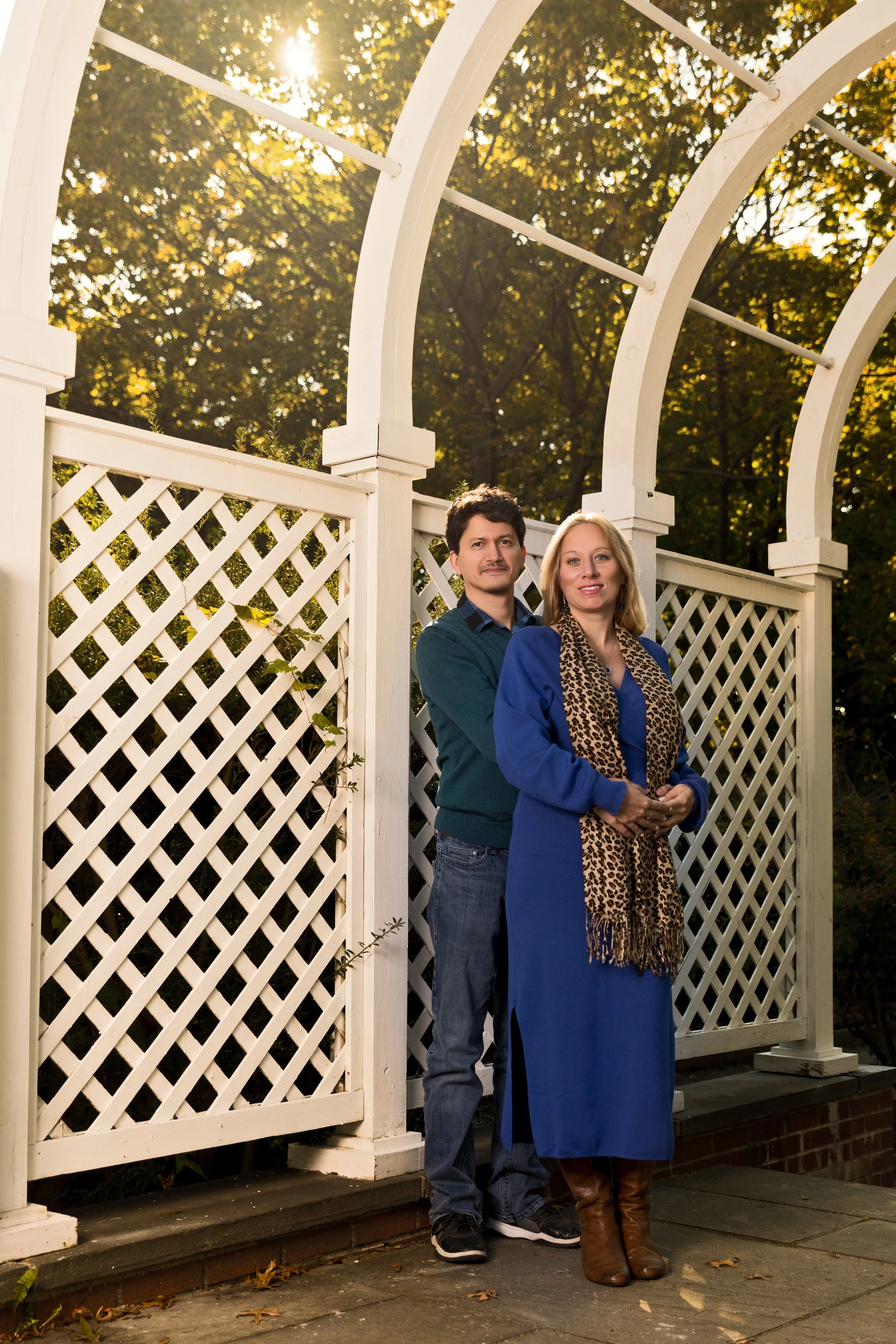 A man and a woman standing in front of a white lattice fence with arches, surrounded by trees in a garden. The woman is wearing a blue dress and a leopard print scarf, while the man is wearing a green sweater and jeans. Sunlight is filtering through 