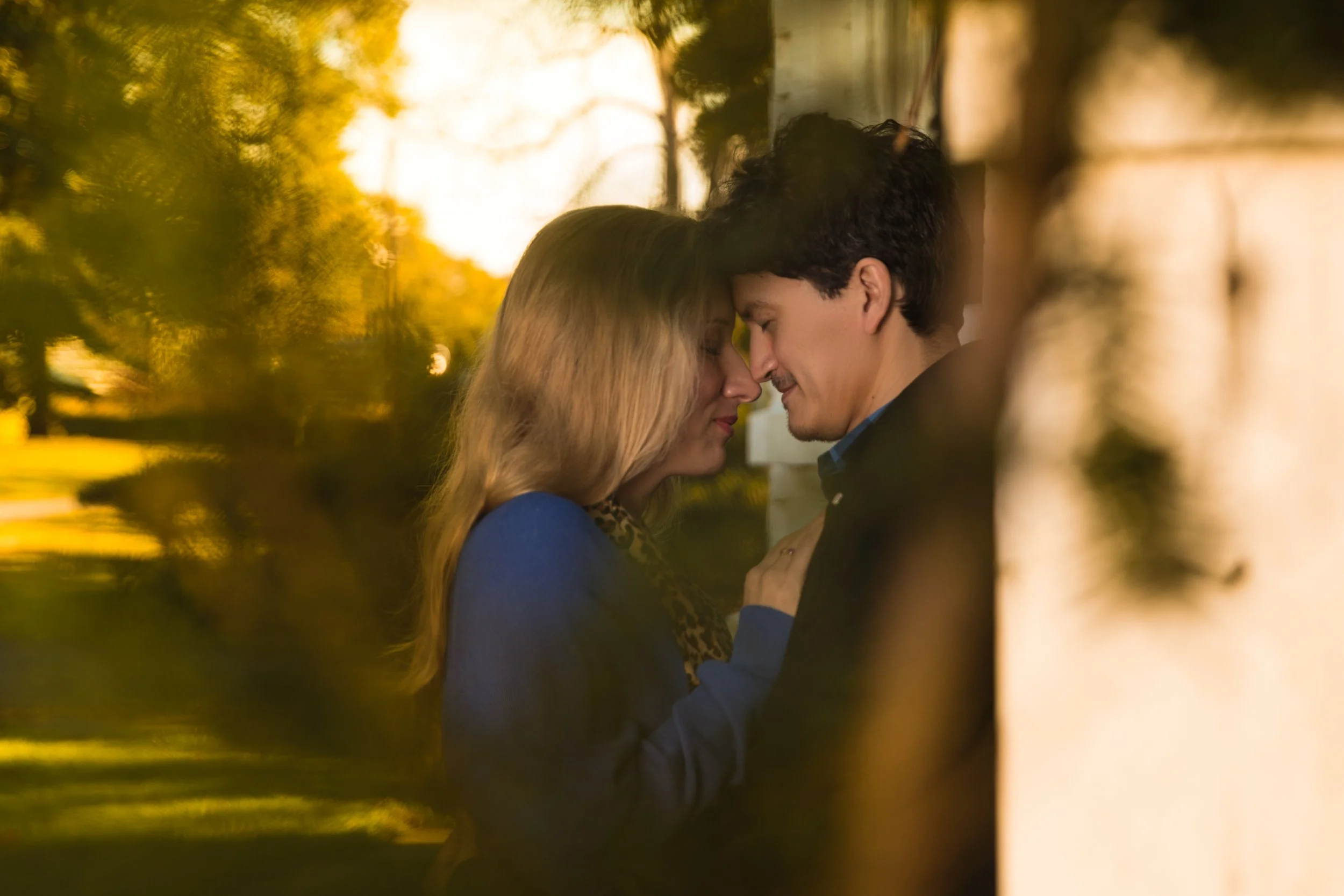 A romantic couple embracing with foreheads touching, surrounded by a blurry outdoor setting.