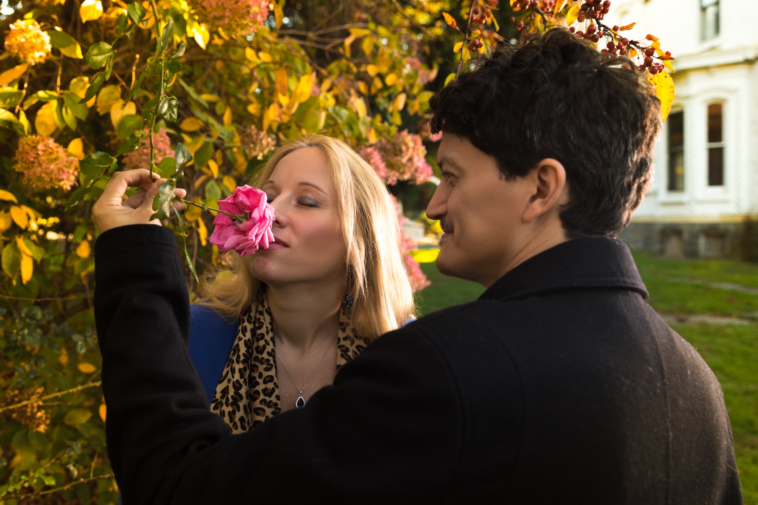 A man holding a pink rose up to a woman's nose, surrounded by colorful autumn leaves.