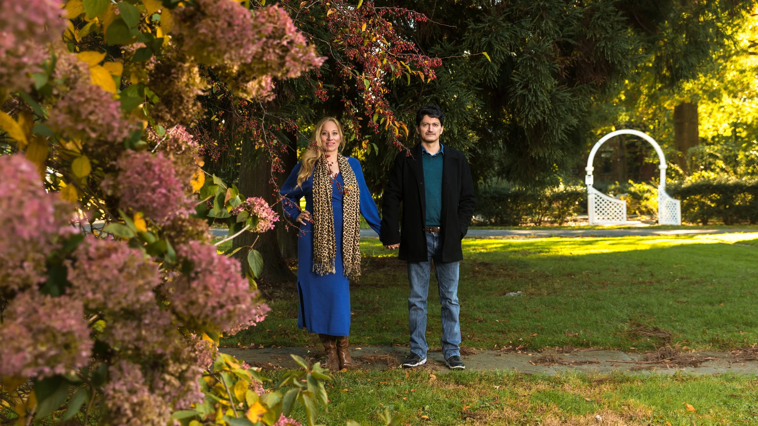 Man and woman standing in park with blooming flowers and a white arbor in the background.