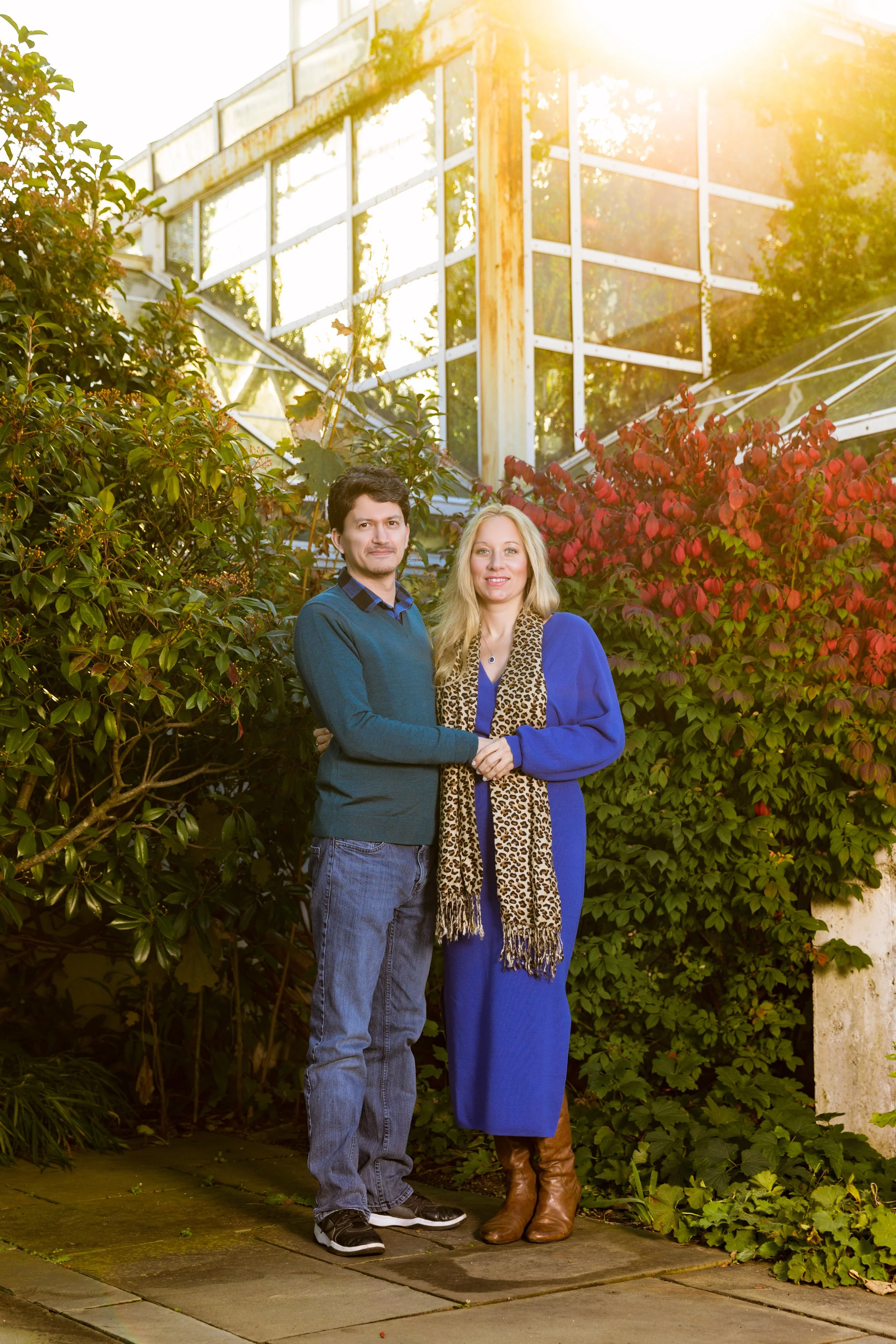 A man and woman standing together in a greenhouse surrounded by green and red foliage, with sunlight shining above. The woman is wearing a blue dress and leopard print scarf, the man is in a blue sweater and jeans.