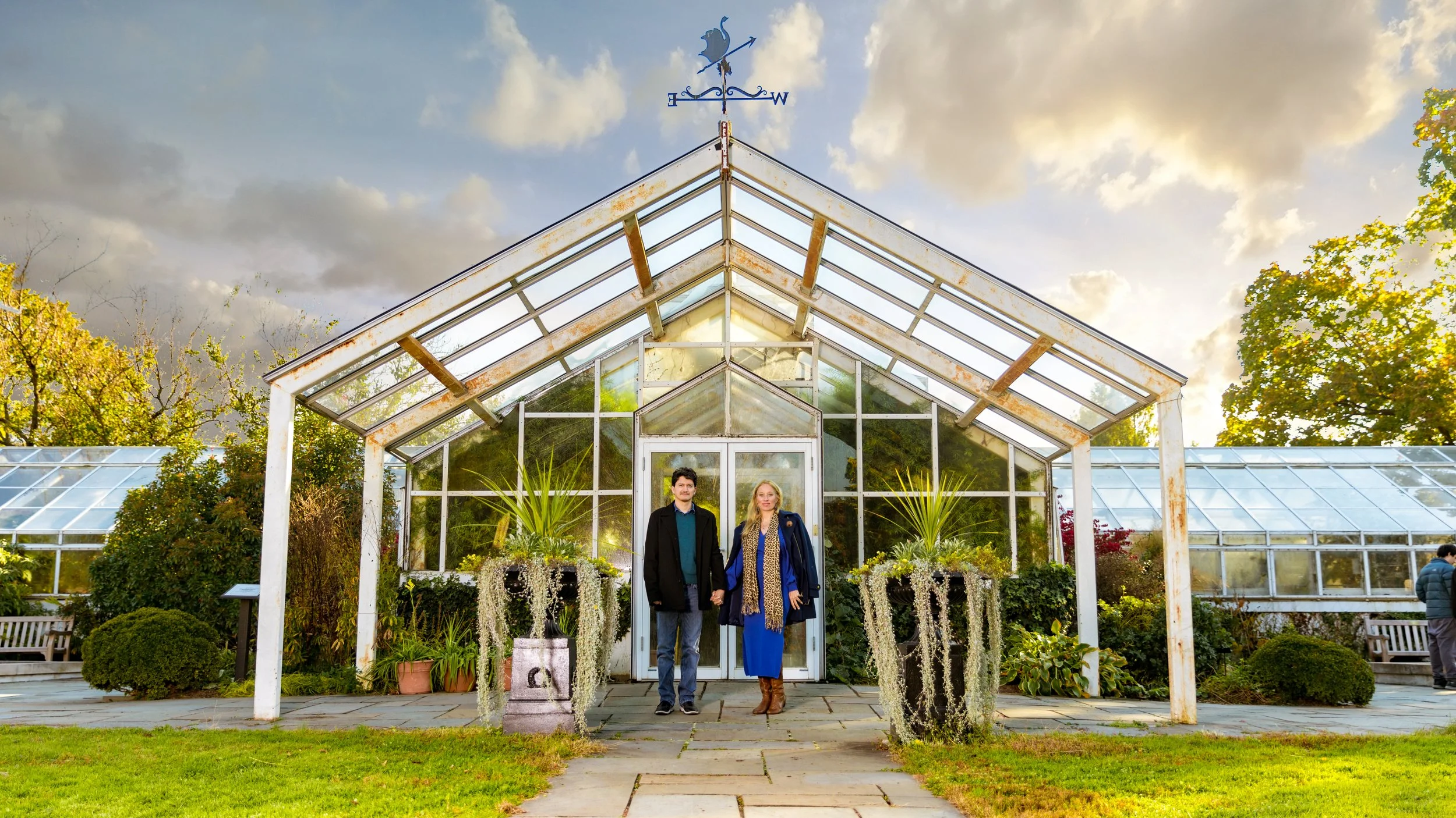 Two people standing in front of a greenhouse with a weather vane on top, surrounded by plants and trees.