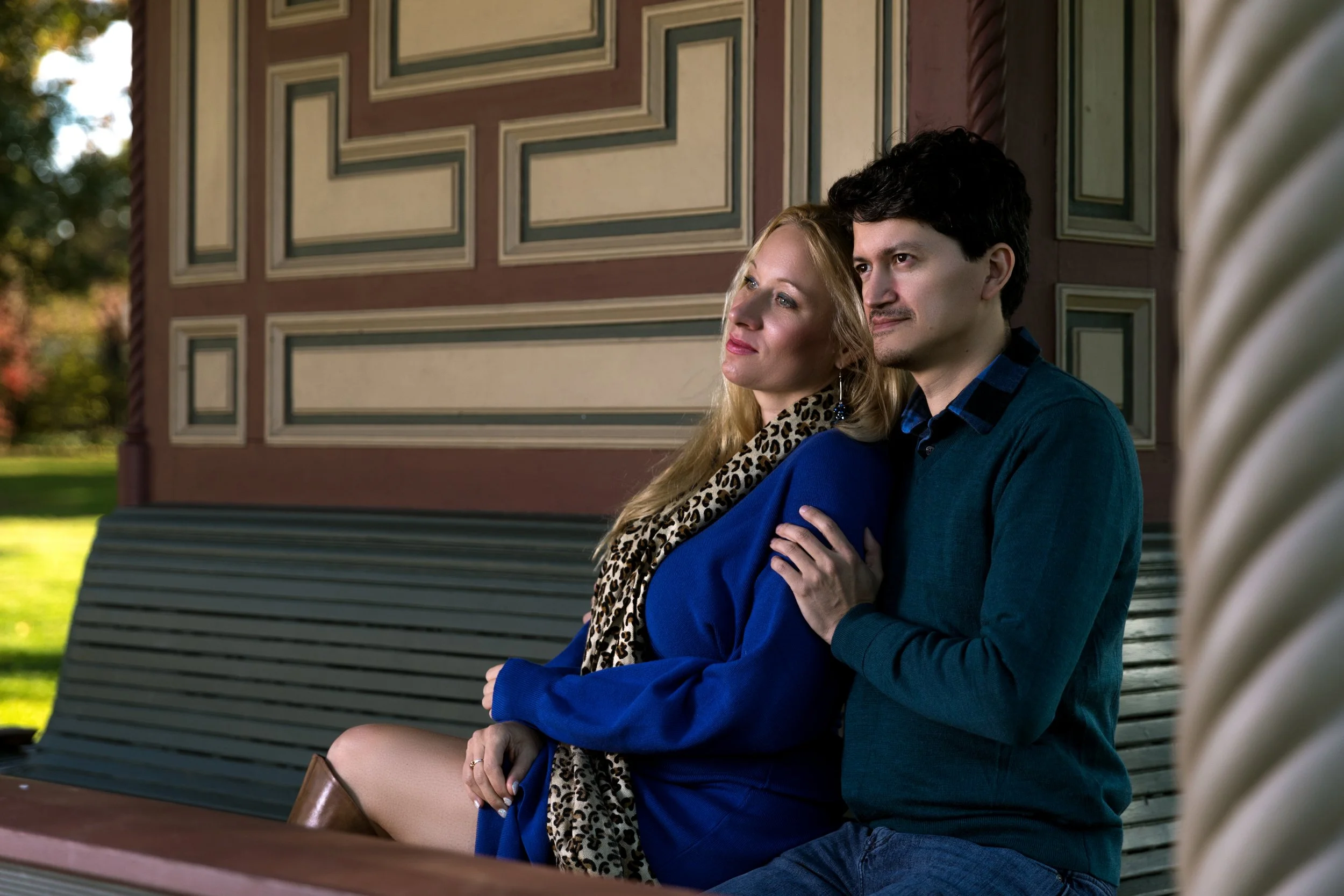 A couple sitting on a bench with geometric patterned walls, woman in blue with a leopard print scarf, man in teal sweater, outdoors.