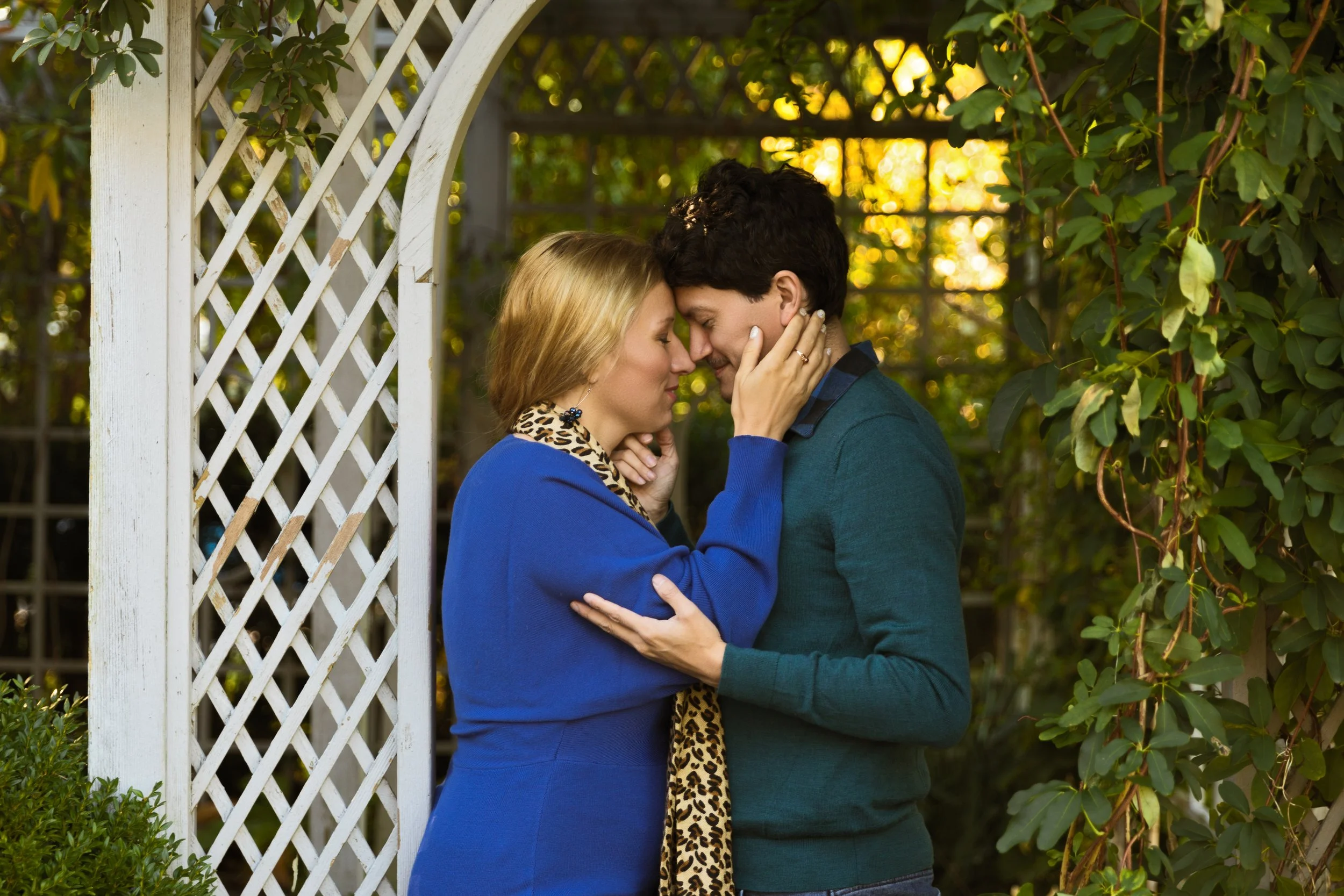 A couple embracing in a garden under a pergola with greenery. The woman is wearing a blue sweater and leopard print scarf, while the man is in a green sweater. They are touching foreheads affectionately.