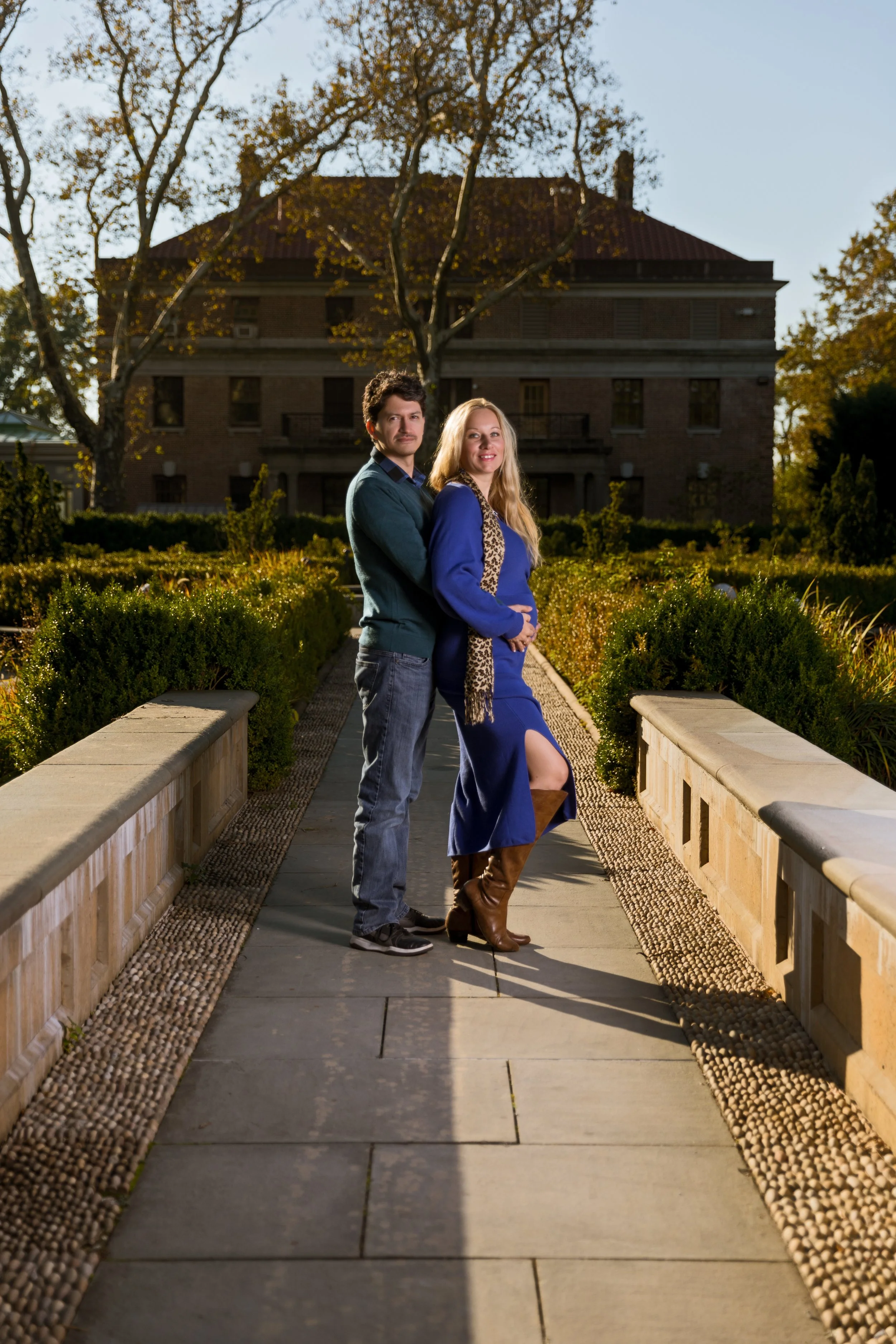 A man and woman standing side by side on a stone pathway in front of a large house. The man is wearing a green sweater and jeans, and the woman is in a blue dress with a scarf and brown boots. The background features trees and a garden.