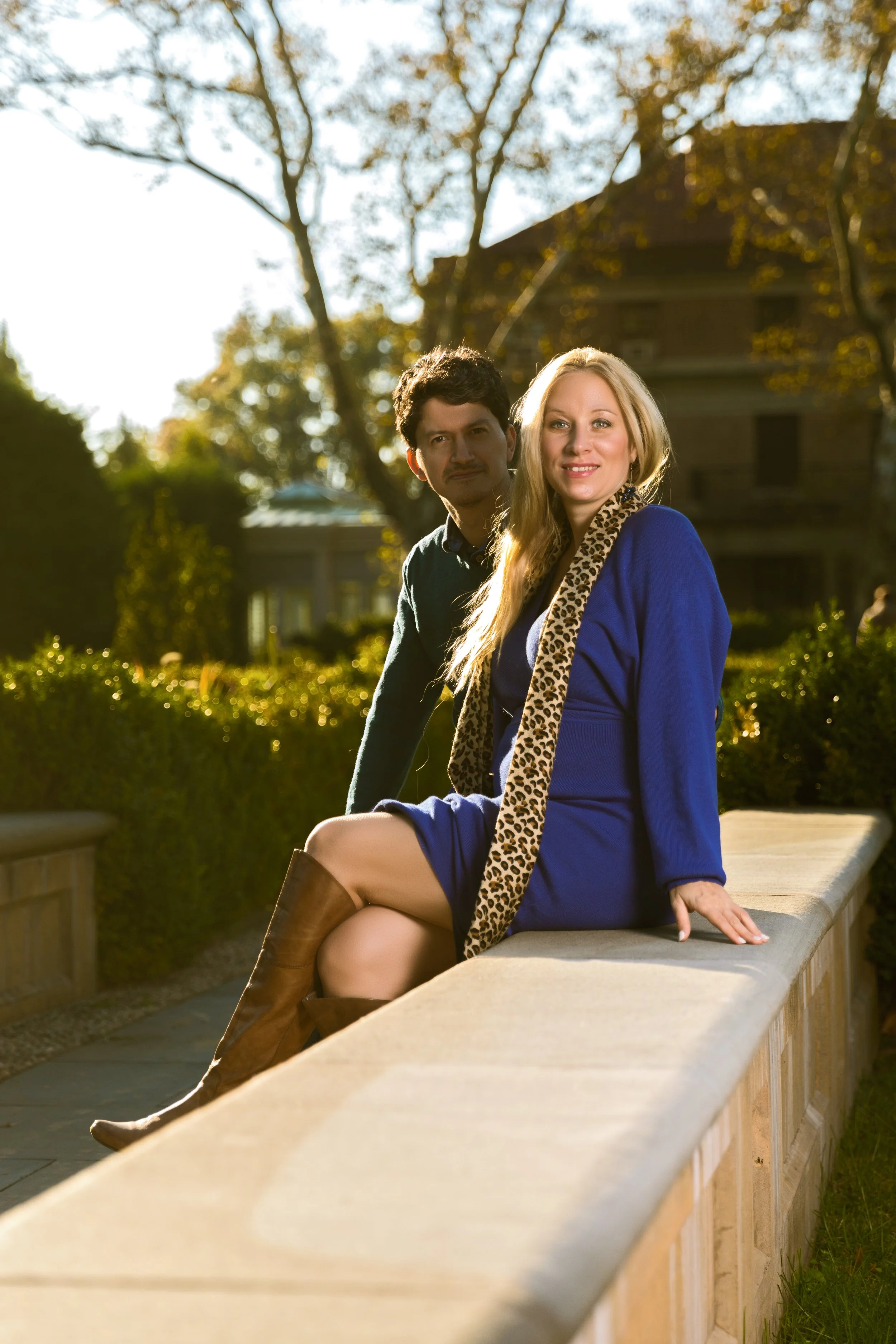 Two people outdoors, one sitting on a stone ledge, wearing a blue dress and brown boots, with a leopard print scarf. The other stands behind in casual attire. Background of trees and buildings.