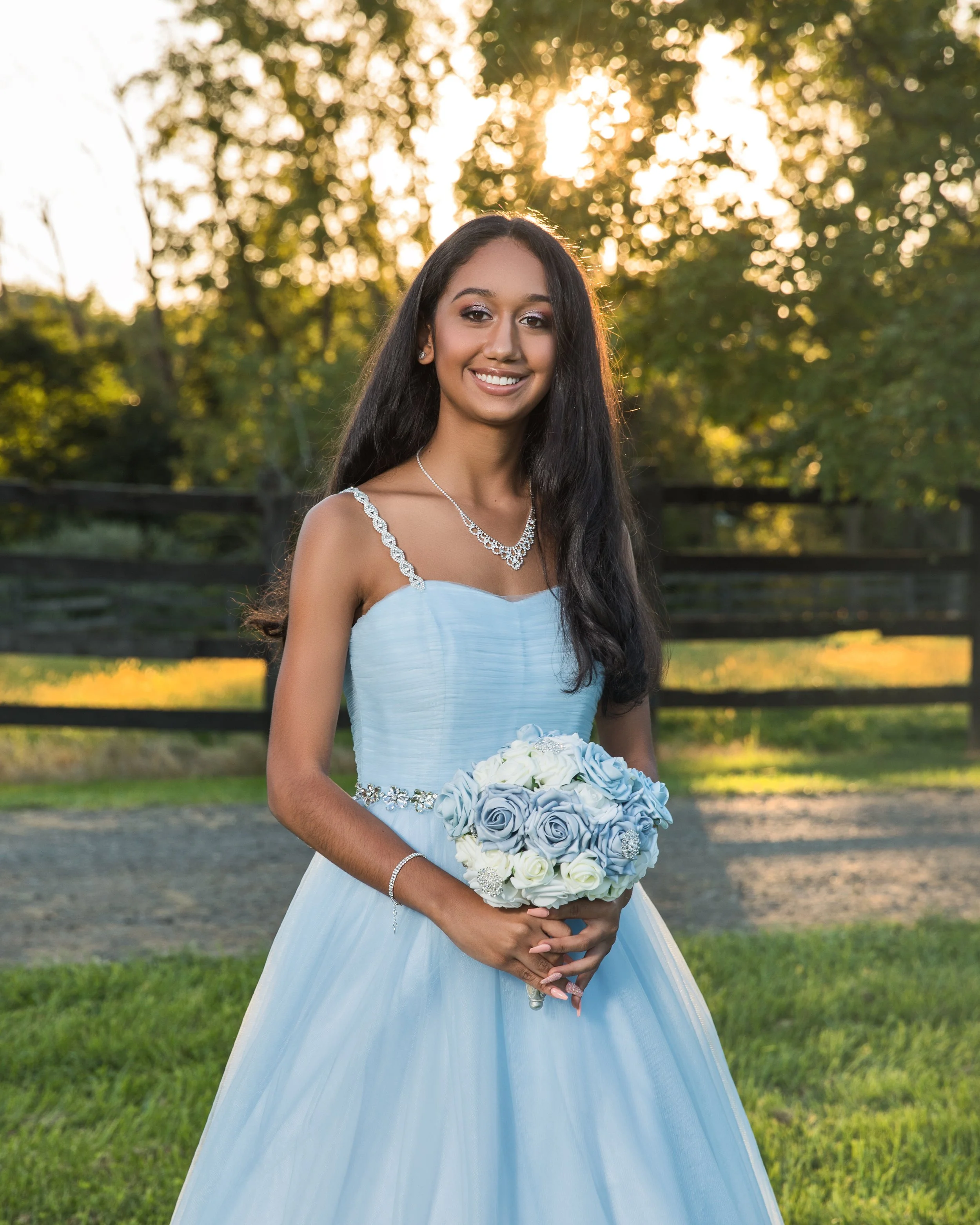 Young woman in a light blue dress holding a bouquet of blue and white roses, standing outdoors with trees in the background.