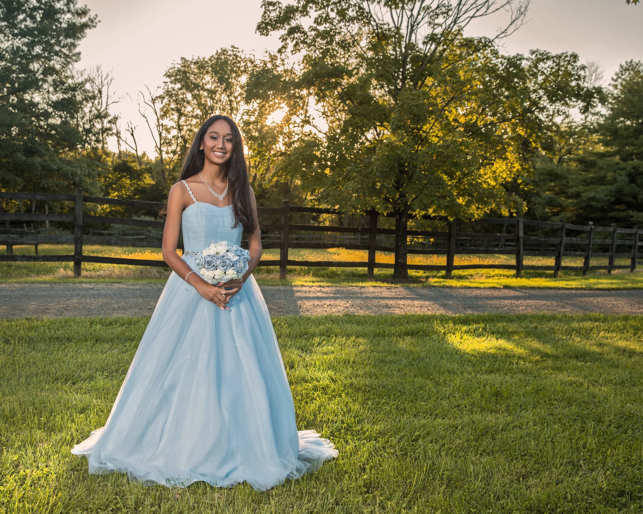 Young woman in a blue gown holding a bouquet, standing on grass with a sunset and trees in the background.