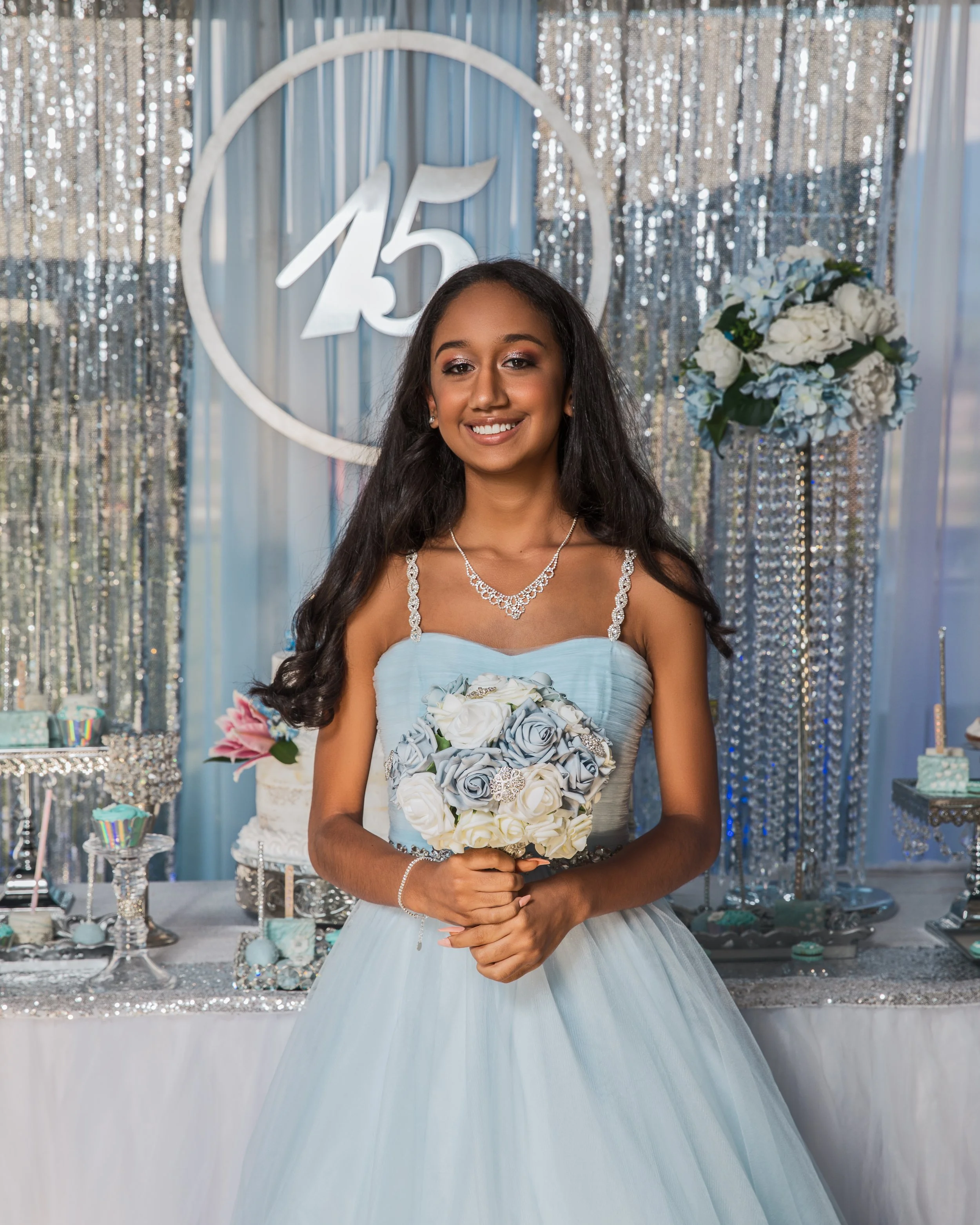 Young girl in a blue quinceañera dress holding a bouquet of roses, standing in front of a decorated backdrop with the number 15 and floral arrangements.
