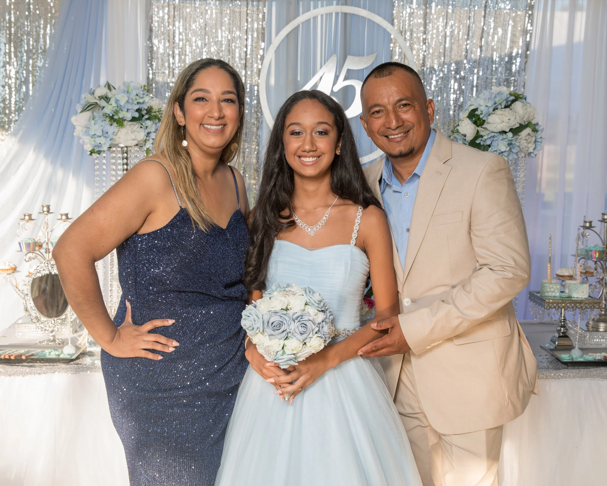 Three people posing at an event with elegant decorations; a young girl in a blue dress holding a bouquet, standing between a woman in a sparkly blue dress and a man in a beige suit. The backdrop includes a '45' sign and floral arrangements.