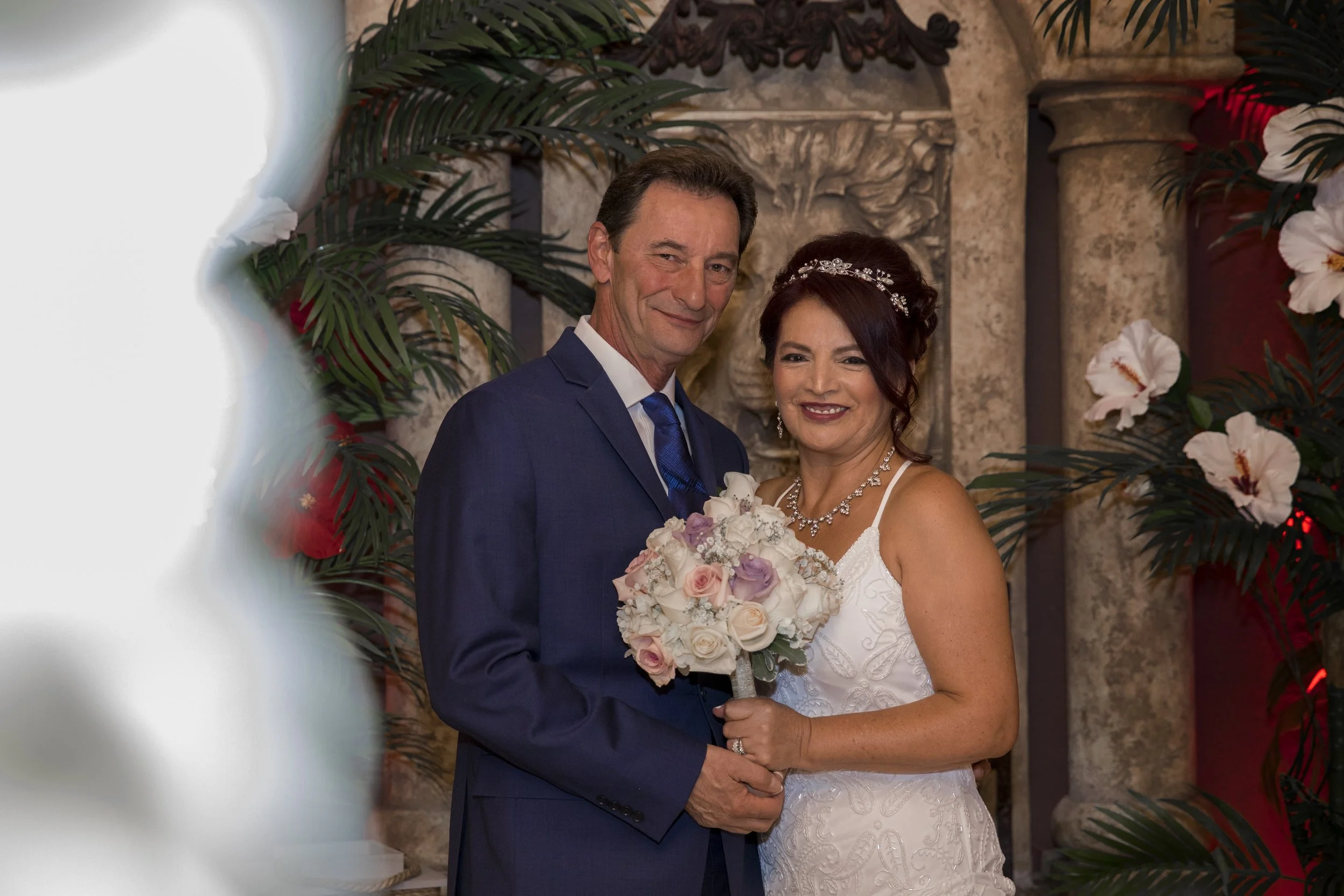 Bride and groom posing with bouquet in front of decorative backdrop, surrounded by plants and floral arrangements.