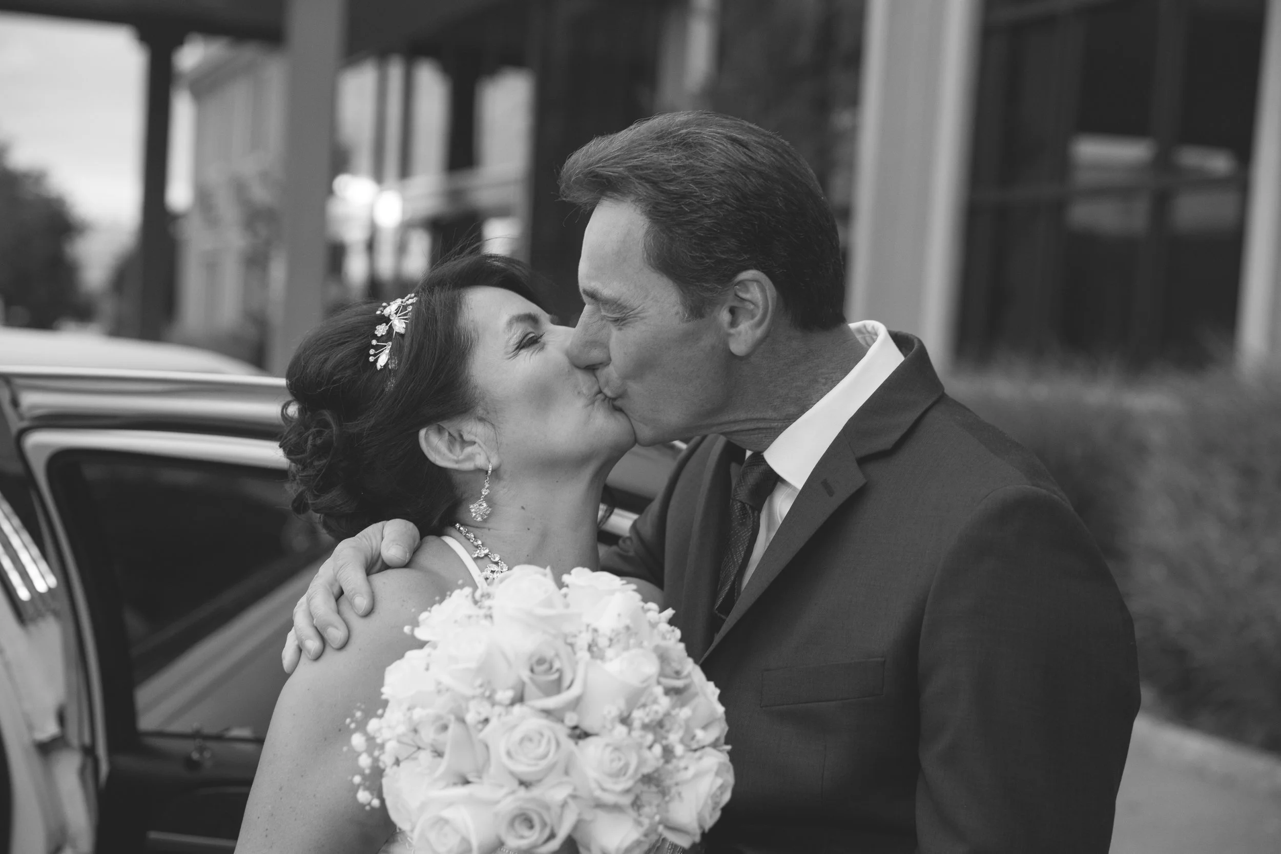 A couple in formal attire kissing outside next to a car. The woman holds a bouquet of roses and wears a hair accessory and earrings. The man is in a suit and tie. Black and white photograph.
