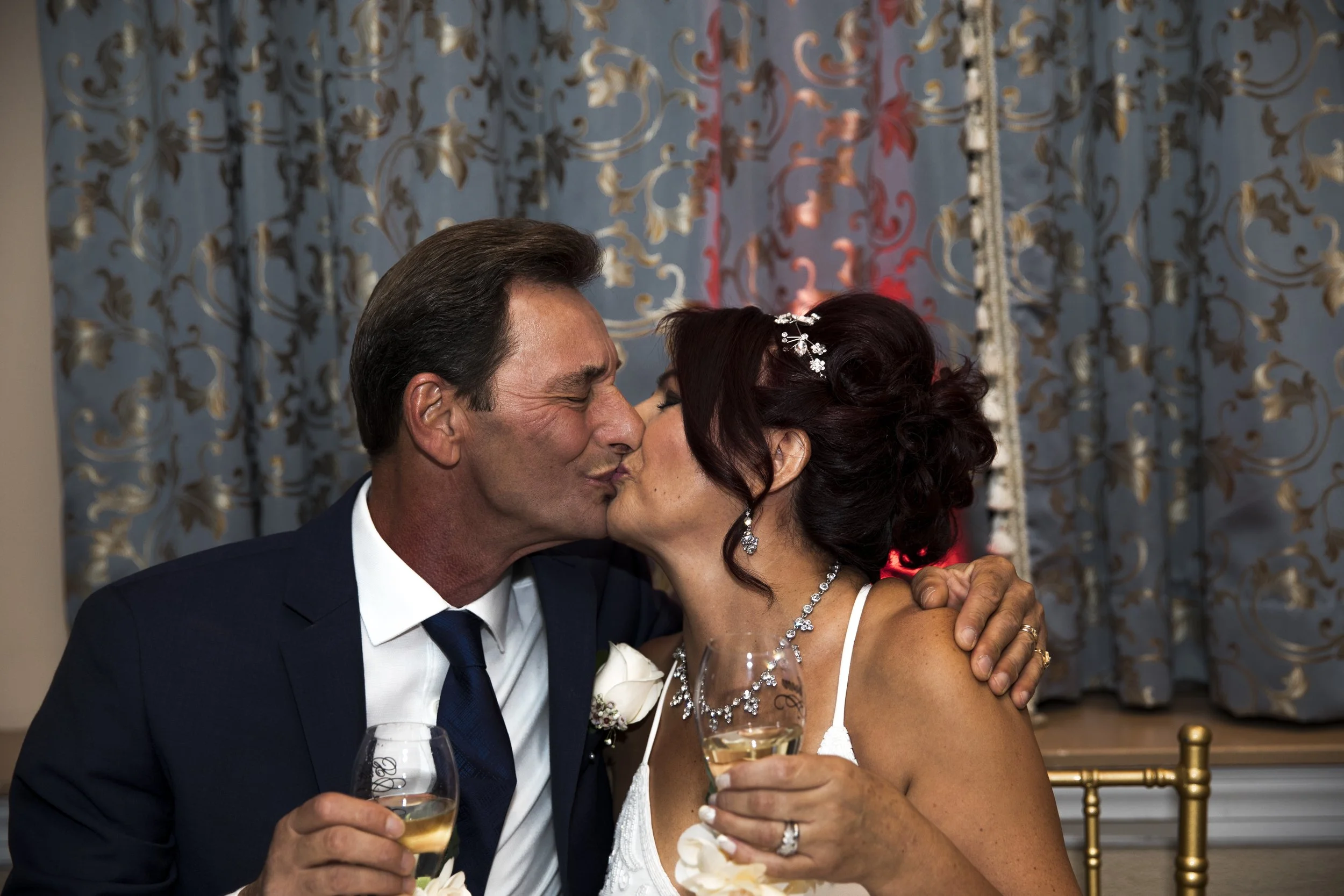 A couple kissing at a wedding, both holding glasses of champagne, with elegant curtains in the background.