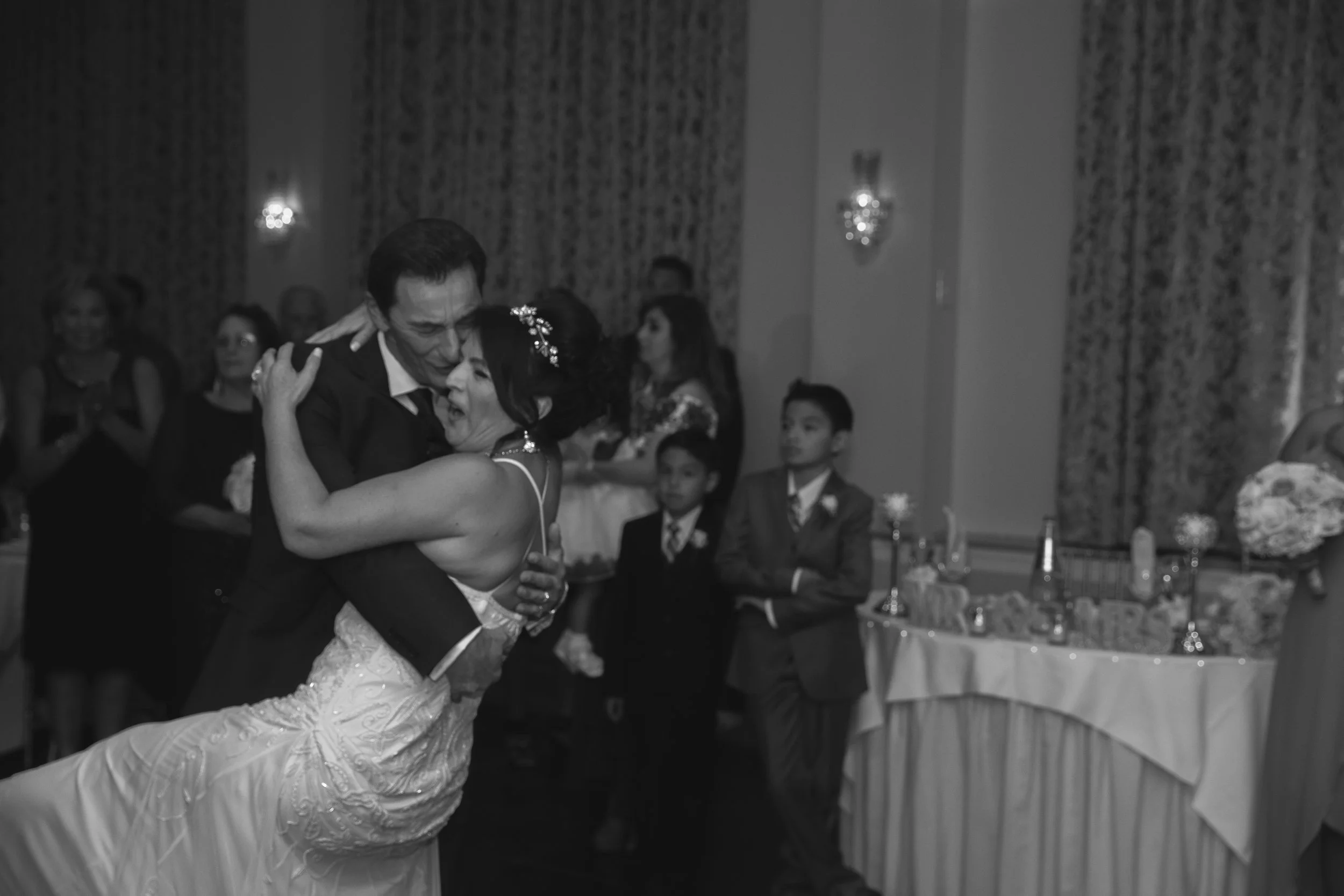 A couple dancing closely at a formal event, with onlookers in the background. The woman is wearing a white dress and the man is in a suit. The atmosphere suggests a celebration, possibly a wedding reception.