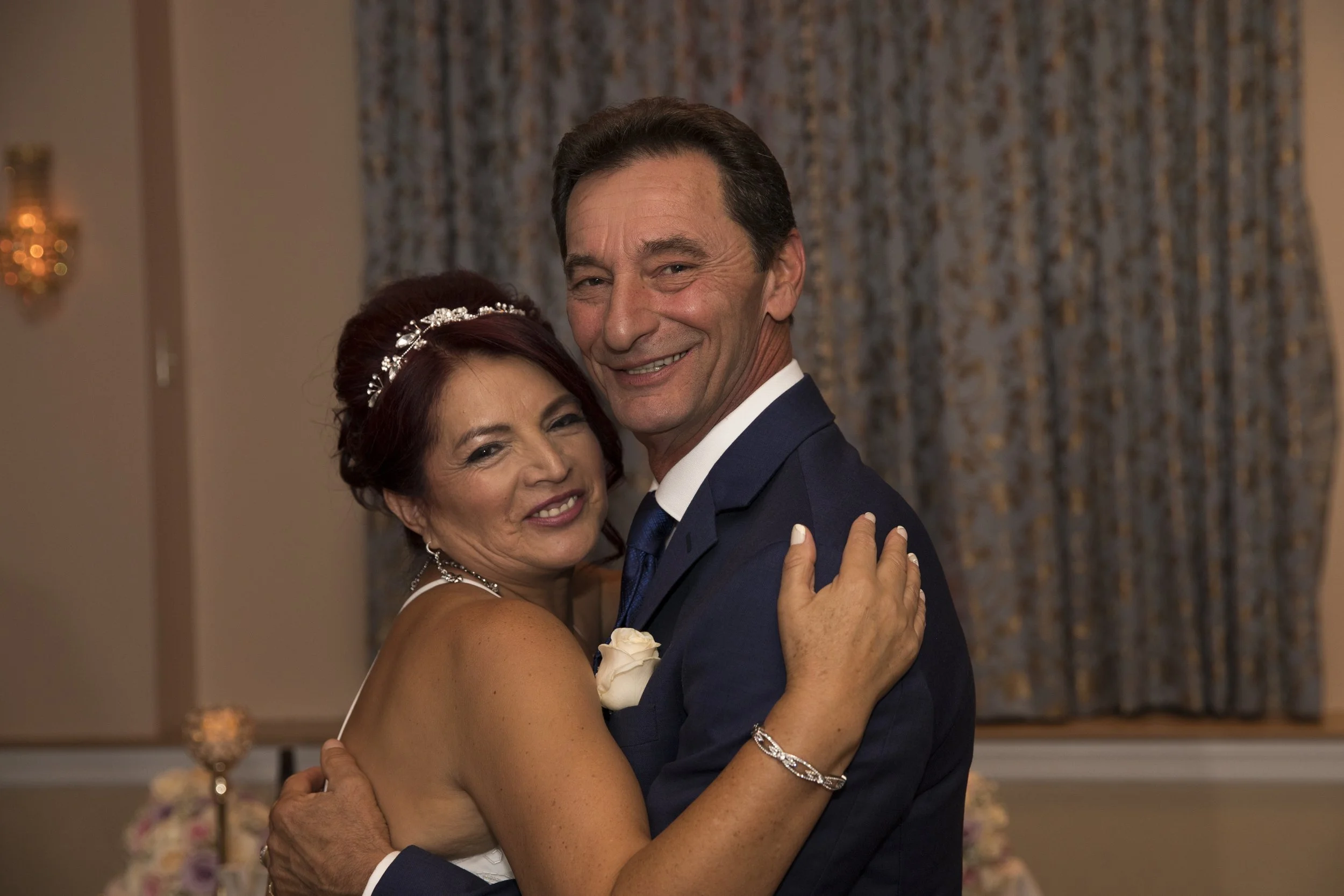 A couple posing happily for a photo at an event. The woman is wearing a white dress with a tiara, and the man is in a blue suit with a boutonniere.