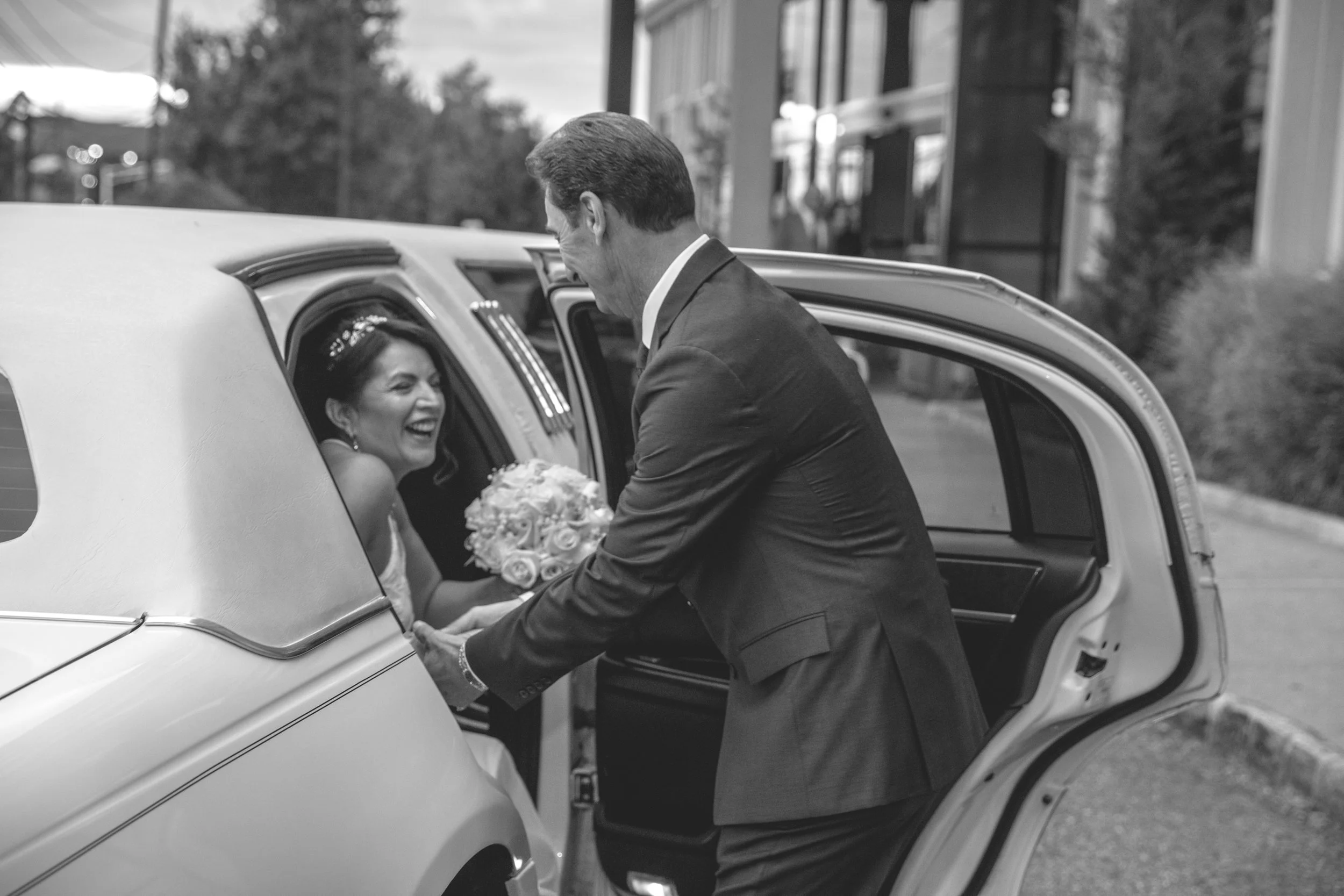 Bride holding bouquet being assisted out of a car by a man, both smiling, in a black and white photograph.