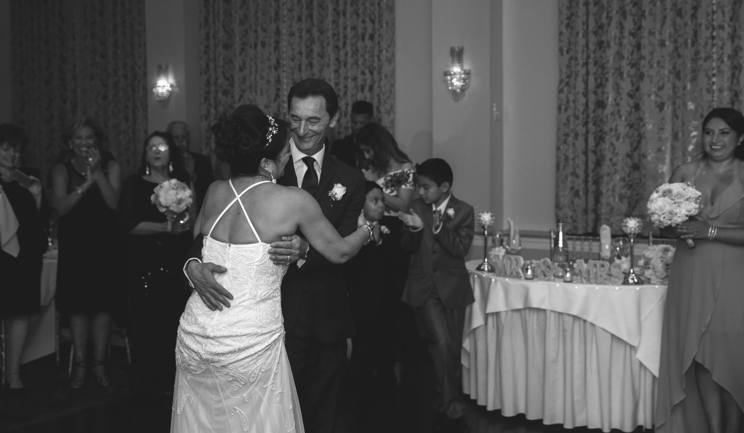 A bride and groom dancing at their wedding reception, surrounded by guests. The bride is wearing a white dress, and the groom is in a suit. Guests, including a child, are clapping and watching the couple dance. Bouquets and decorations are visible in