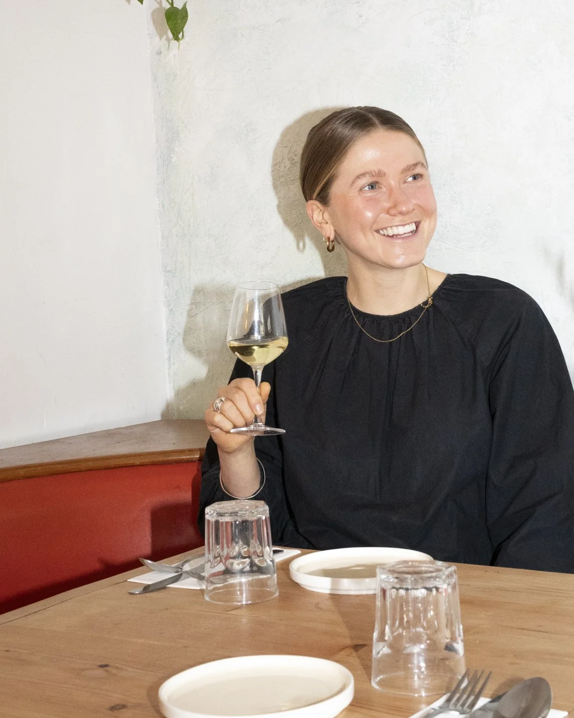 A woman with short hair, wearing a black top and gold jewelry, is sitting at a table with a glass of white wine in her hand. She is smiling and looking to her right in a well-lit, casual setting.