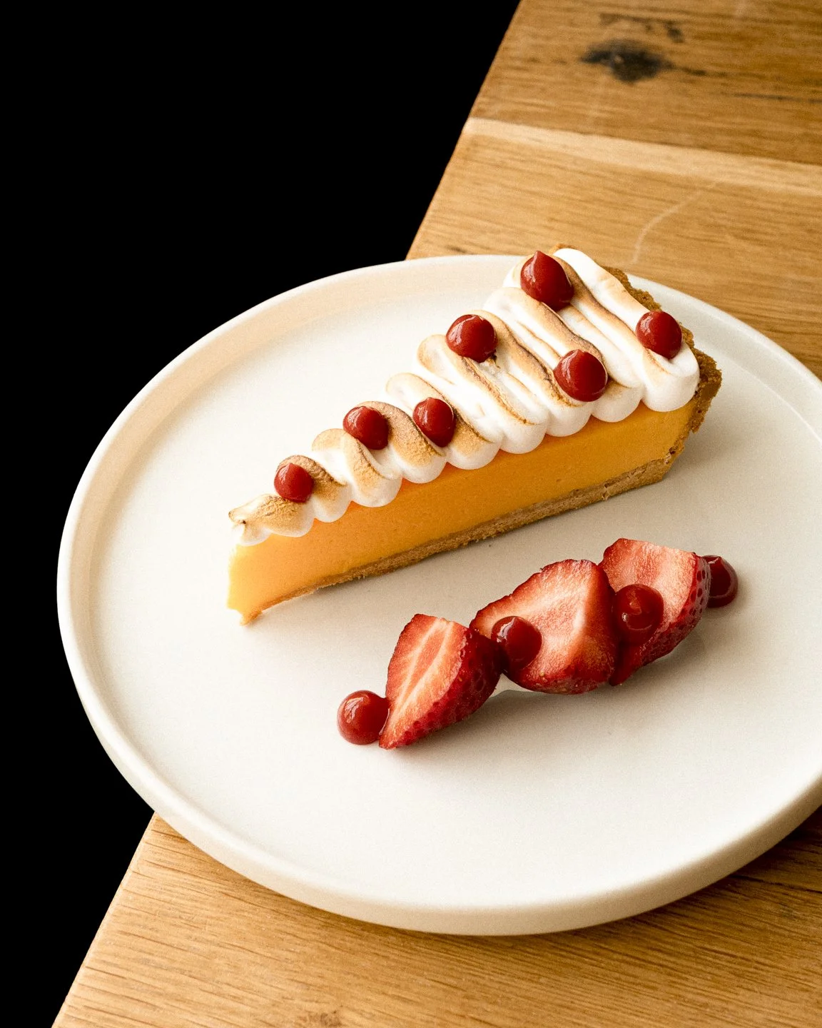 A slice of lemon tart garnished with whipped cream and red berries, served with sliced strawberries and berries on a white plate.