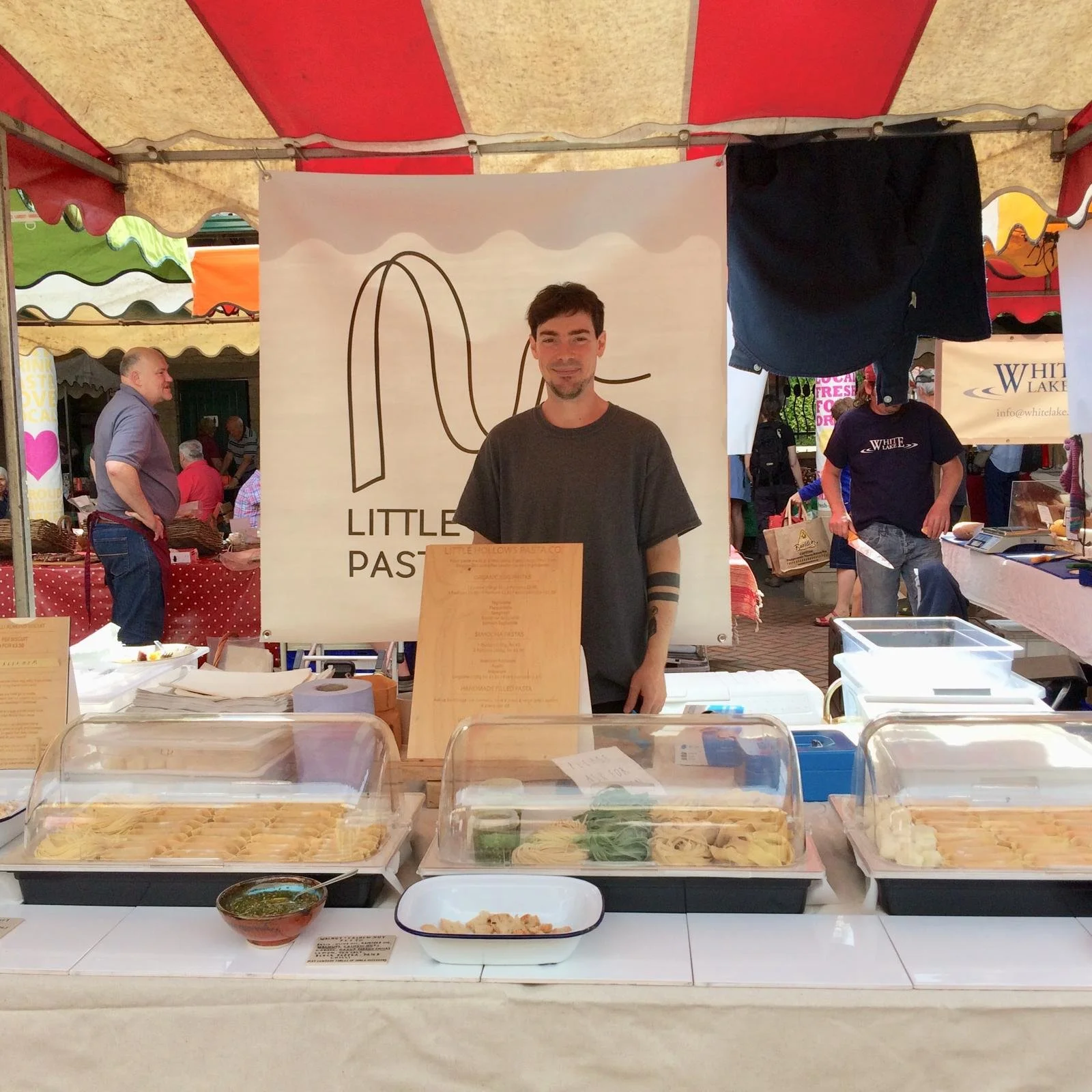 A man stands behind a table at a food stall selling pasta, with containers of cooked pasta in front of him and a sign that reads 'Little Hollow's Pasta Co.' in the background. The scene is set at an outdoor market with colorful tents and people in the background.
