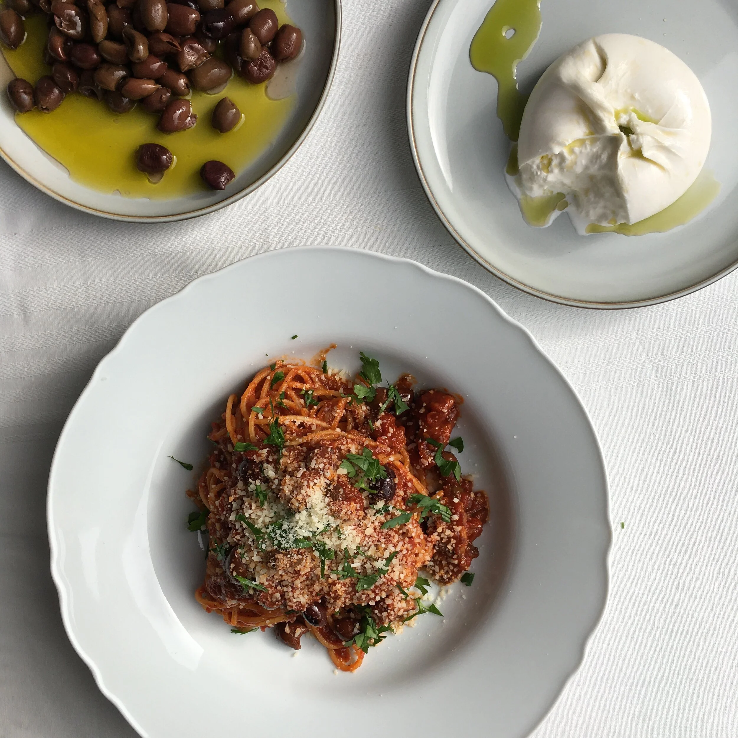 A plate of spaghetti with tomato sauce, garnished with chopped parsley and grated cheese, served on a white dish. Another plate contains a ball of mozzarella cheese drizzled with olive oil. A third plate has Kalamata olives in olive oil, also served on a white dish. The dishes are placed on a white tablecloth.