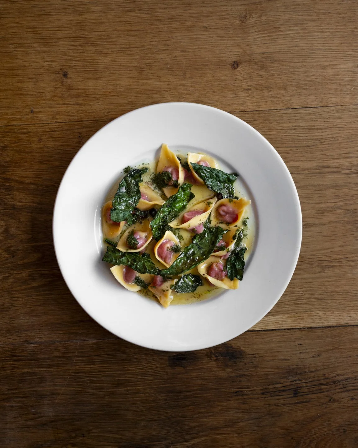 Plate of tortellini pasta with green pesto, spinach leaves, and slices of ham on a wooden table.