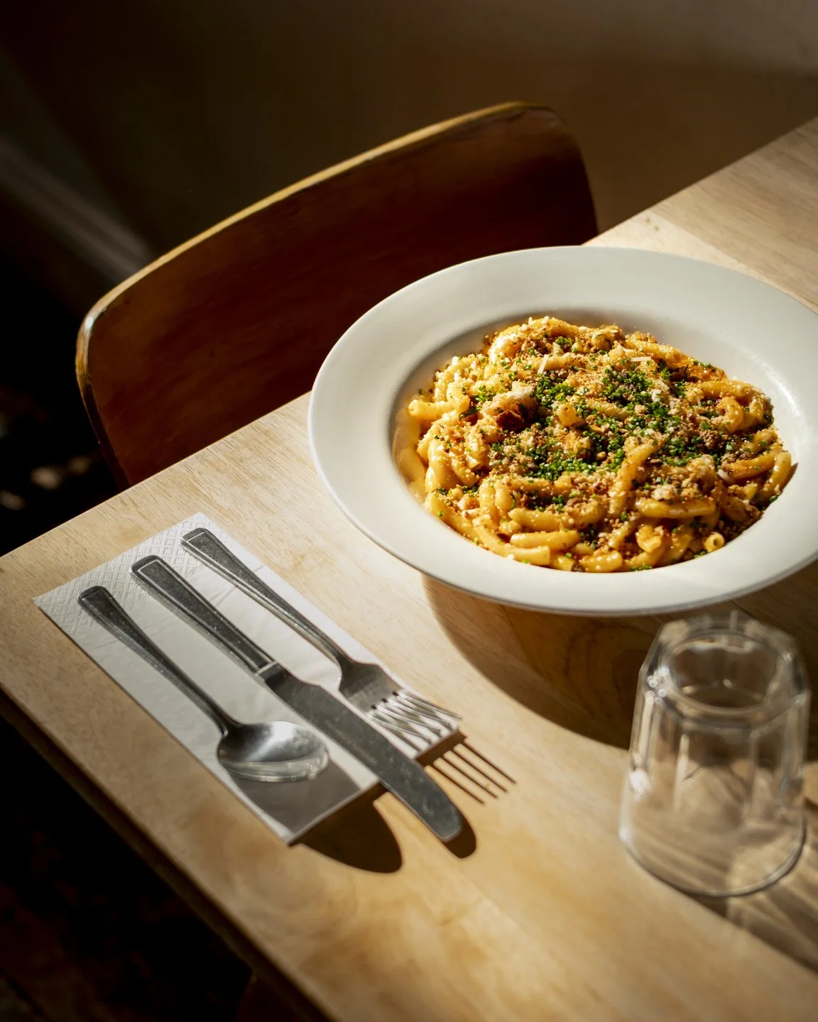 A white bowl filled with a pasta dish, likely Bolognese, topped with grated cheese and chopped herbs, sits on a wooden table next to a napkin with a fork and spoon, with a glass of water nearby.