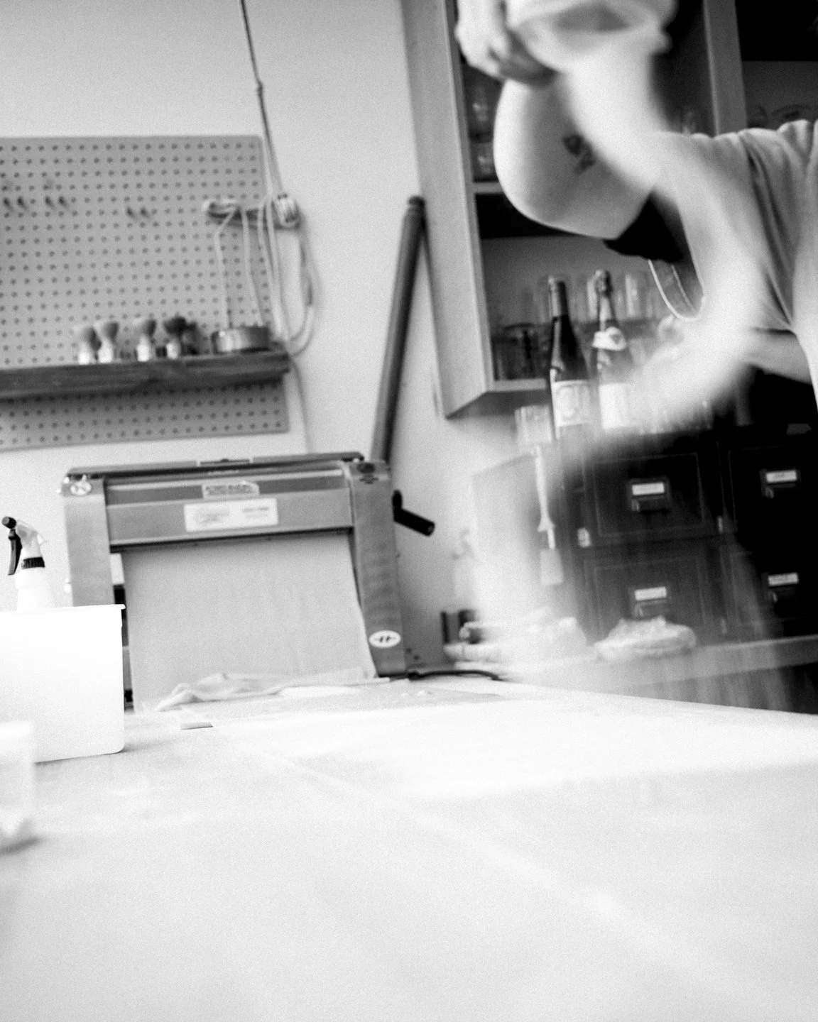 A person working in a workshop or kitchen, wearing a mask, with shelves and a machine in the background.
