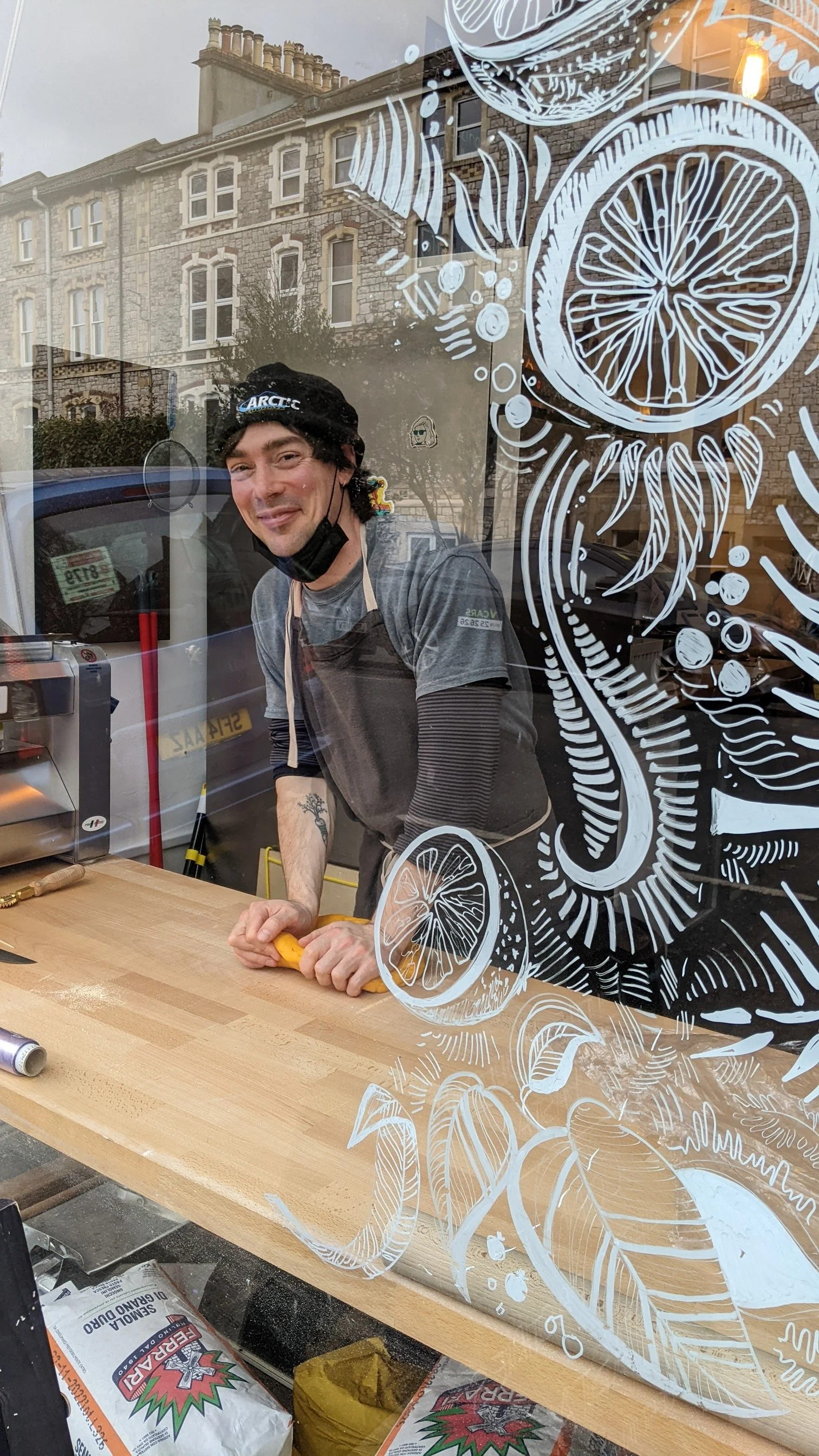 A man wearing a black beanie, a gray T-shirt, and a face mask around his chin, is peeling a yellow bell pepper in a kitchen. The photo is taken through a window with white hand-drawn citrus fruit and leaf illustrations.