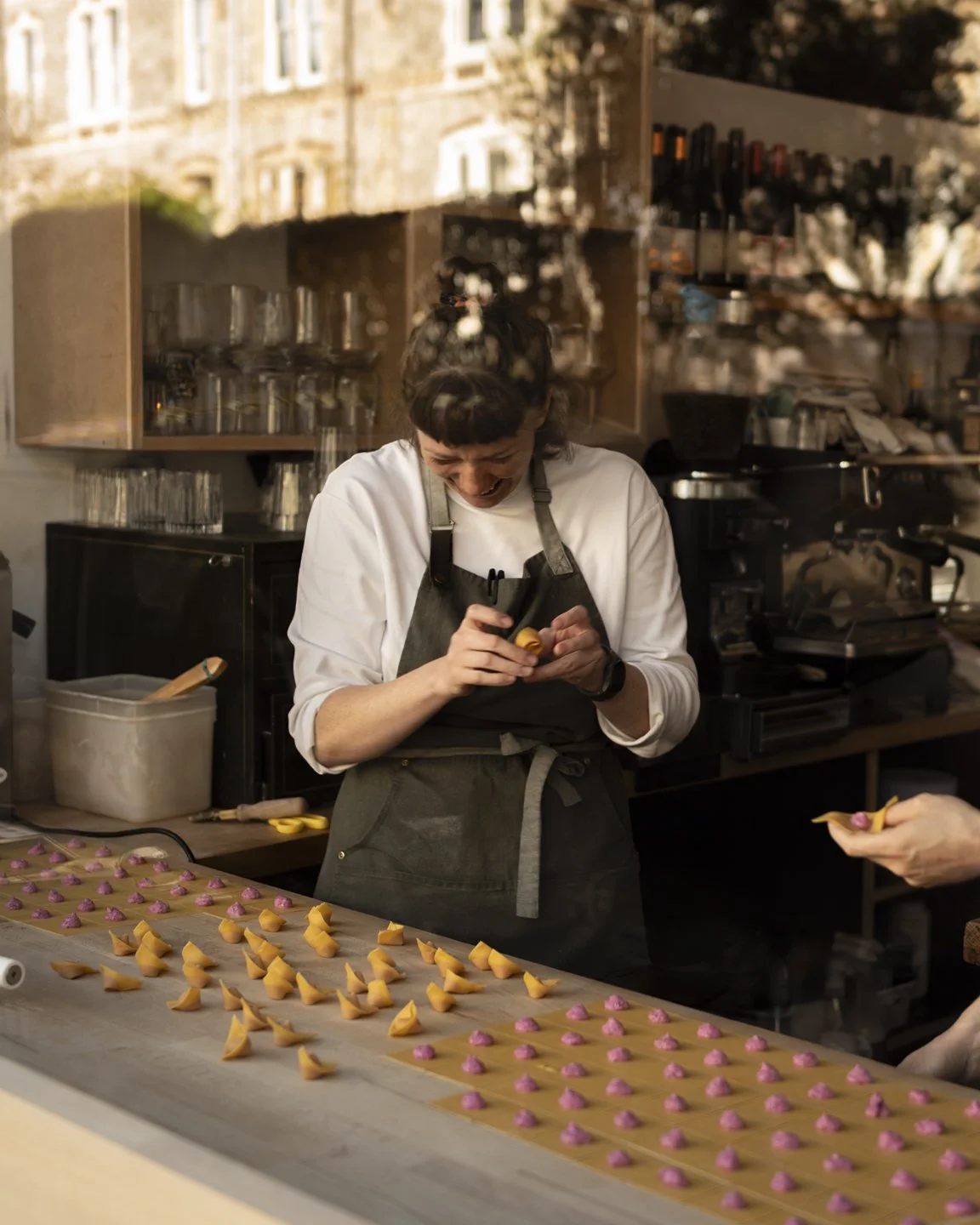 A woman in a black apron is working in a kitchen, smiling as she prepares colorful pastry bundles on a tray, with a person handing her a piece.