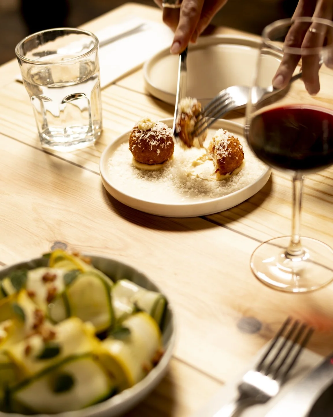 A dining scene featuring a plate of fried cheese balls on a bed of grated cheese, a glass of water, a glass of red wine, and a bowl of rolled zucchini slices topped with herbs on a wooden table.