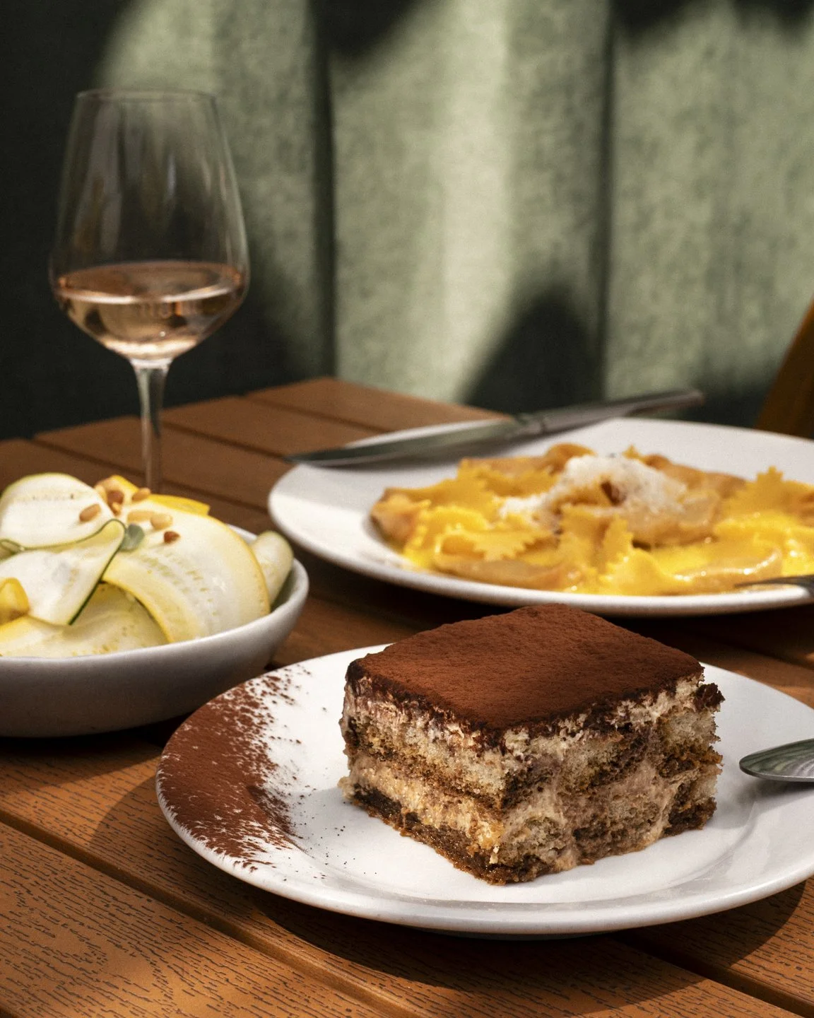 A dessert plate with tiramisu, a bowl of sliced pears with pine nuts, a glass of rosé wine, and a plate of ravioli with cheese and sauce on a wooden table.