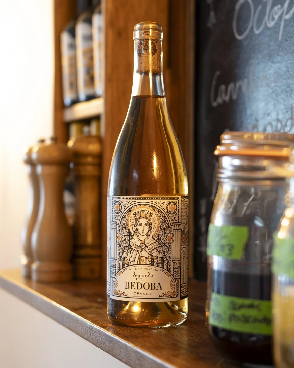 A glass bottle of orange wine with a label featuring a detailed illustration of a crowned woman holding a staff, sitting on a throne, against a wooden background with jars and containers nearby.