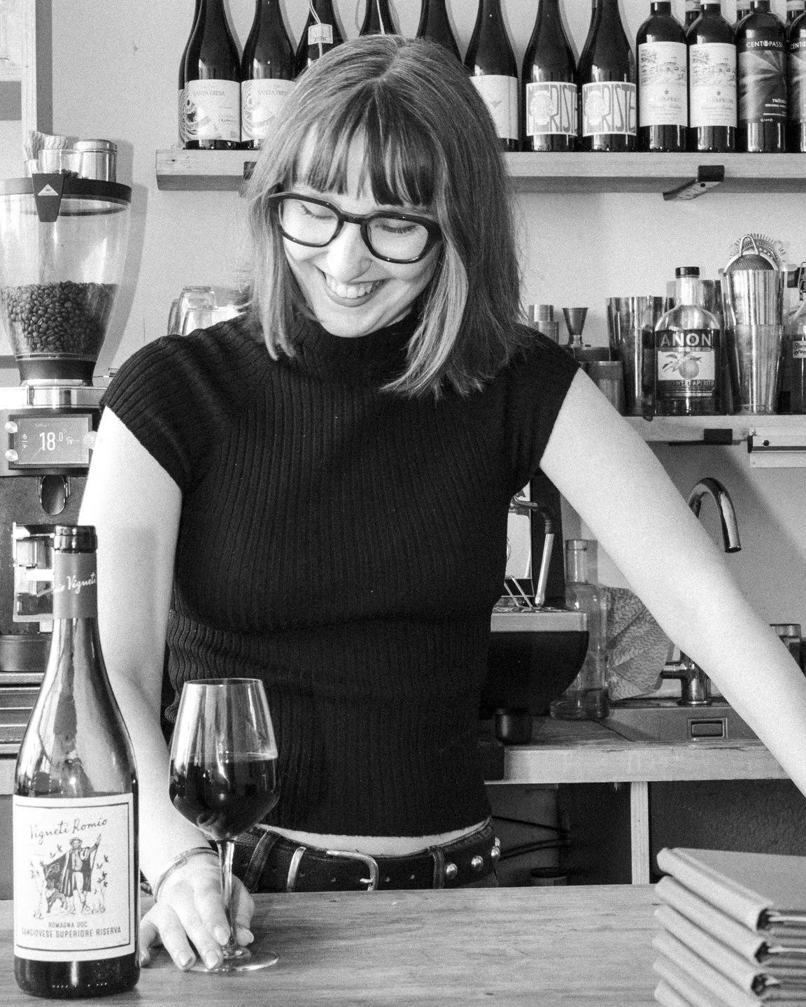 A woman with glasses in a black sleeveless top smiles while standing behind a counter with a wine glass and a bottle of red wine in a cafe or bar setting.