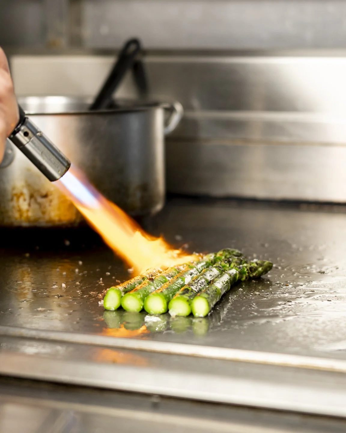Green asparagus spears being charred on a stovetop with a blowtorch flame