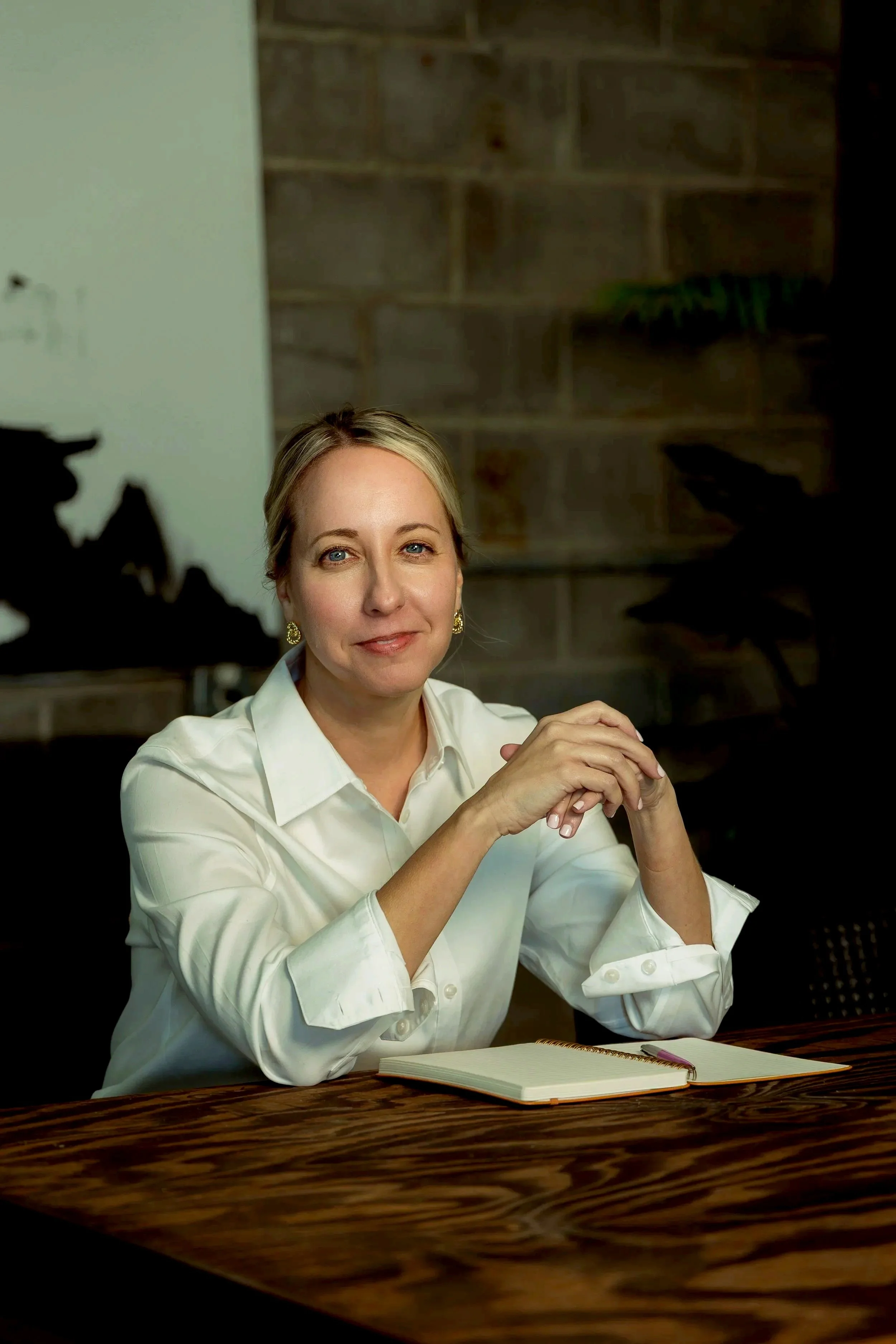 A woman with blonde hair and blue eyes, wearing a white button-up shirt and gold earrings, sitting at a wooden table with a notebook and pen, smiling softly in a room with a brick wall and plant in the background.