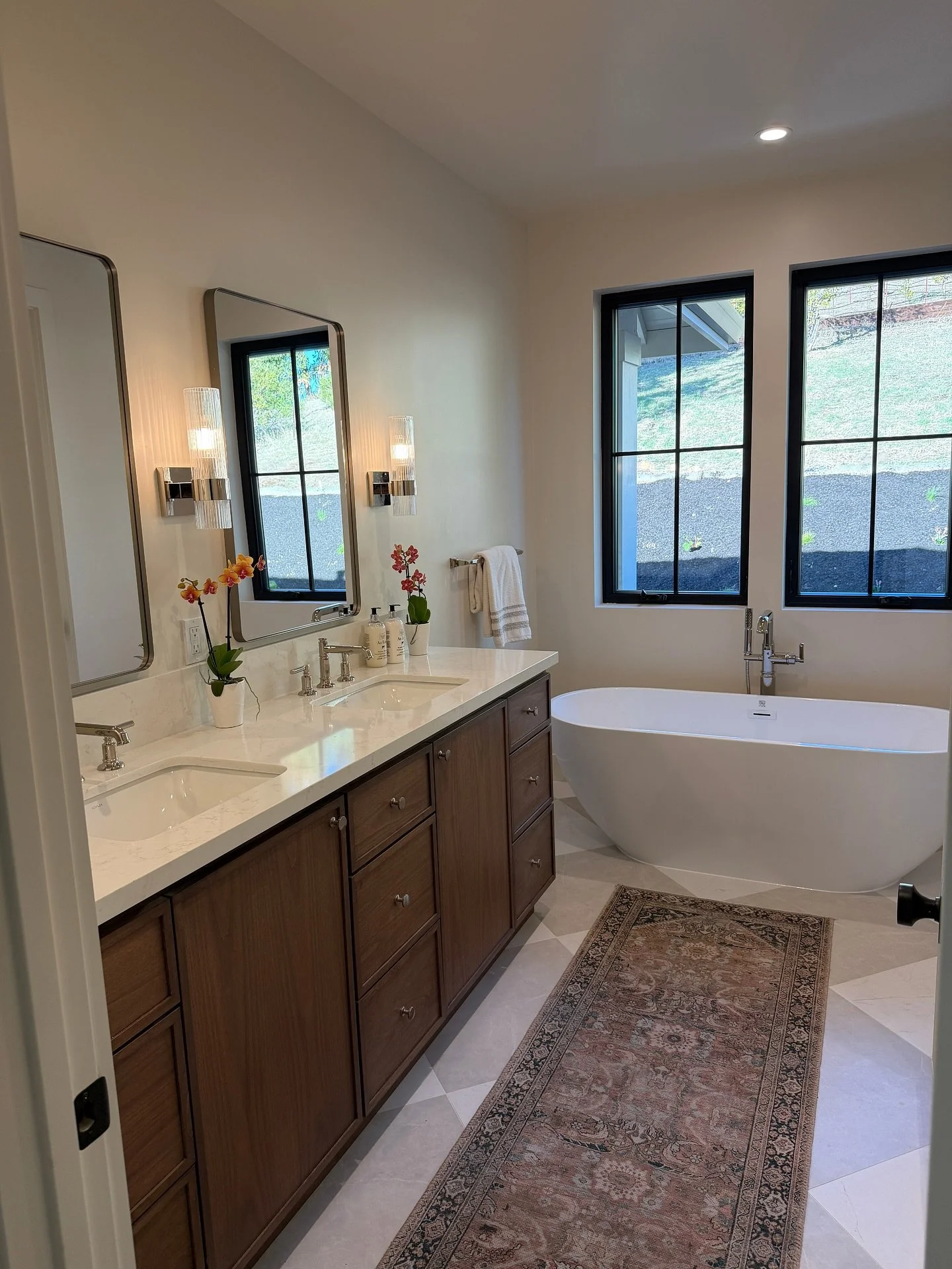 Calm is a design choice.

In the primary bath, we paired natural wood cabinetry with polished nickel and a soft stone floor &mdash; creating a palette that feels quiet but elevated.

A freestanding tub anchors the space, surrounded by filtered mornin