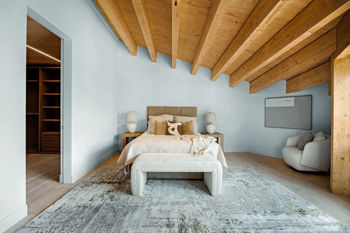 O House bedroom staged by MKOD with layered cream bedding, a textured bench and exposed timber ceiling