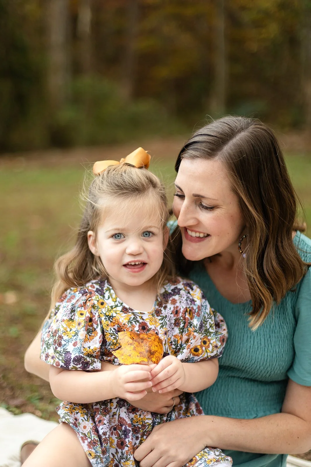 A woman and young girl smiling outdoors in a park, with the girl holding a yellow autumn leaf.