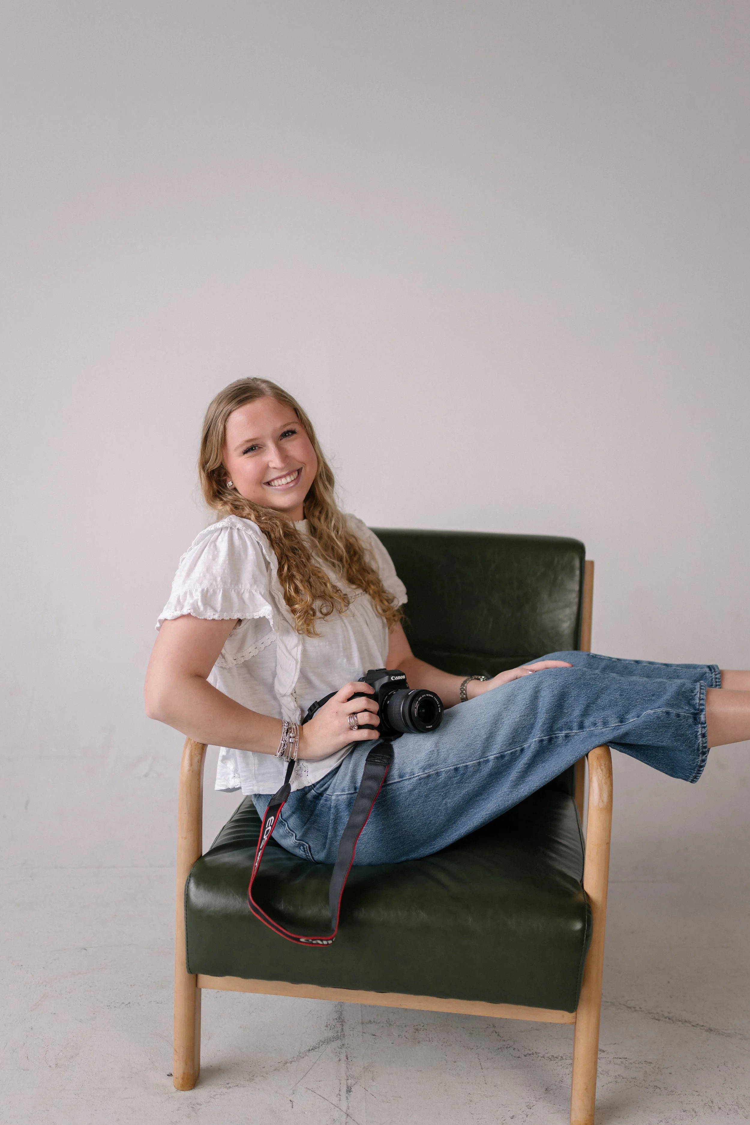 Chloe Williams, creative assistant at Bright & Gray Creative, smiling in a studio portrait seated casually in a green leather chair holding a Canon camera, wearing a white eyelet top and wide-leg jeans, representing photography and content production