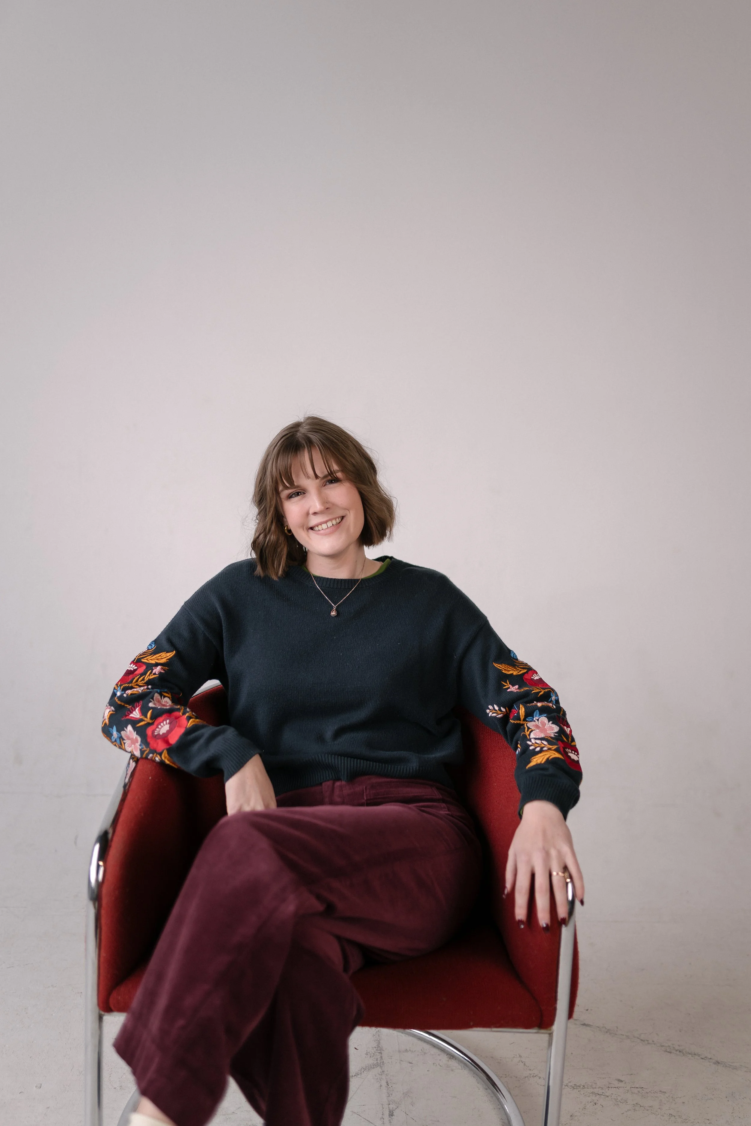 Lakyn Phillips, design coordinator at Bright & Gray Creative, smiling in a studio portrait seated in a red chair wearing a dark teal sweater with floral sleeves and burgundy corduroy pants, representing creative design services for small businesses a