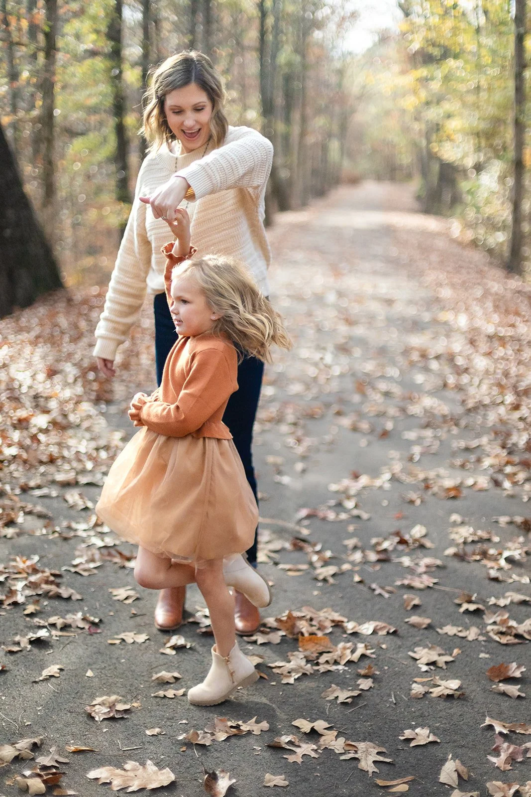 A woman and a young girl are playing together on a fallen leaf-covered path in a wooded area during autumn. The woman is holding the girl's hand and laughing, while the girl is mid-spin, wearing a tan skirt, a rust-colored top, and beige boots.