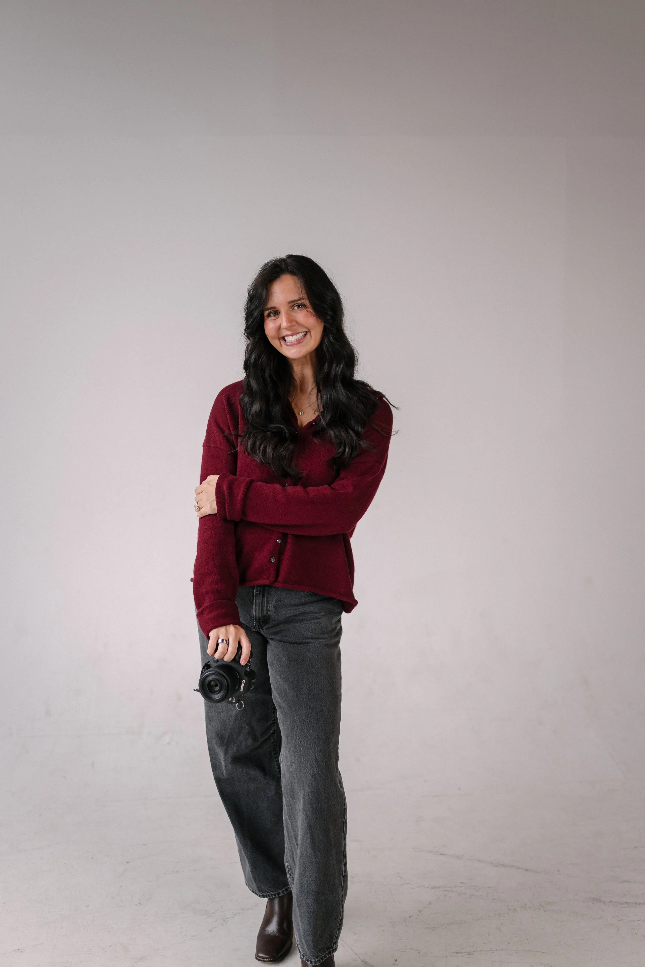 Morgan Brown, design assistant at Bright & Gray Creative in Birmingham, AL, smiling in a studio portrait holding a camera and wearing a burgundy cardigan and gray wide-leg jeans, representing creative design and photography services for small busines
