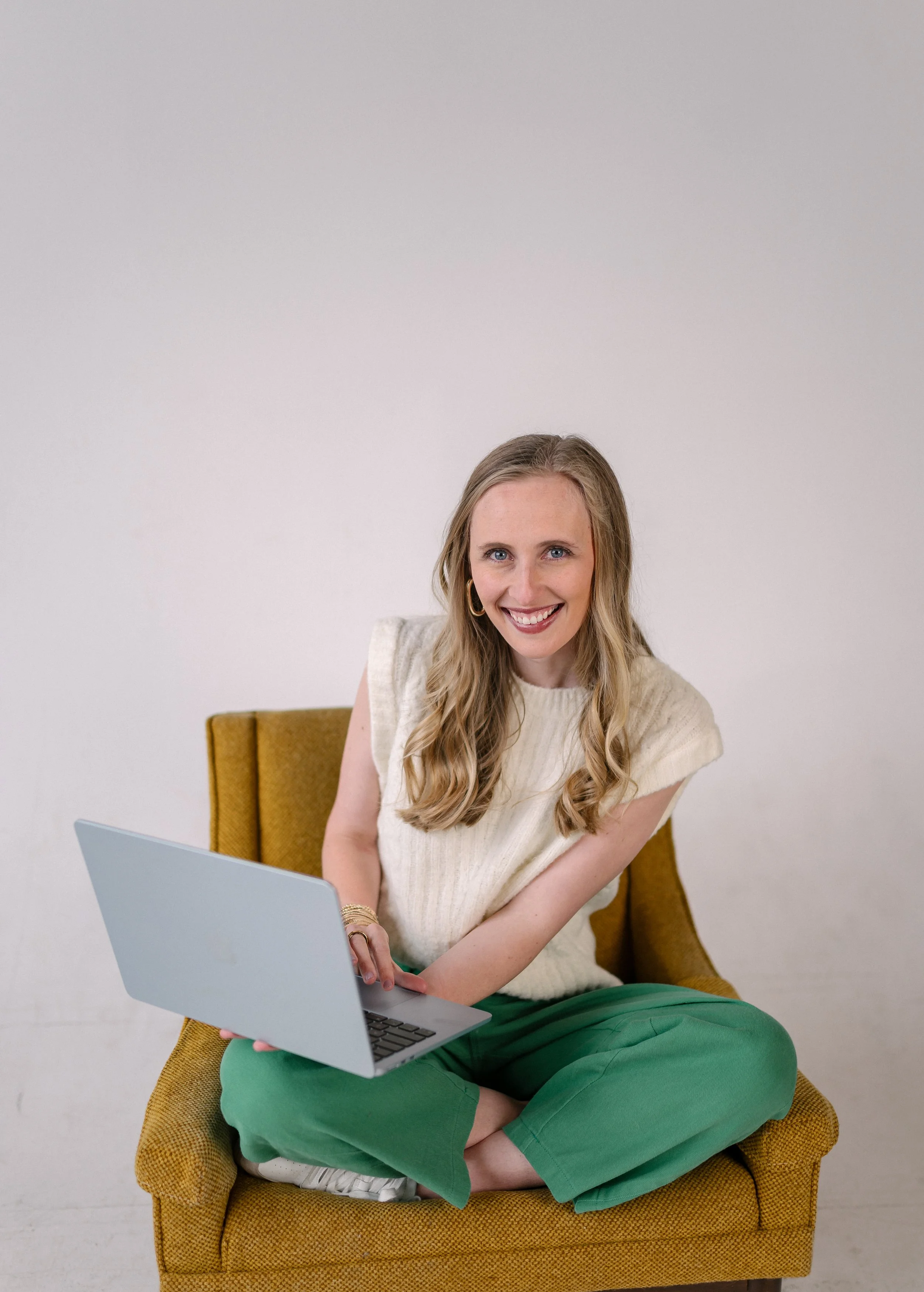 Ella Hillman, project coordinator at Bright & Gray Creative in Birmingham, AL, smiling while seated in a mustard yellow chair with a laptop, wearing a cream sweater top and green wide-leg pants, representing creative project management and marketing 