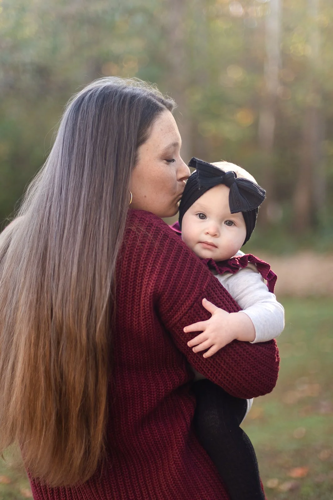 A woman with long brown hair holding and kissing a baby girl with a black bow headband in an outdoor setting with trees in the background.